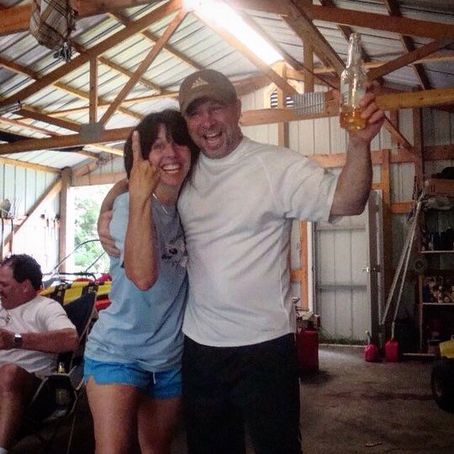 Smiling couple posing arm-in-arm inside a wooden shed; the man raises a beer bottle in celebration.