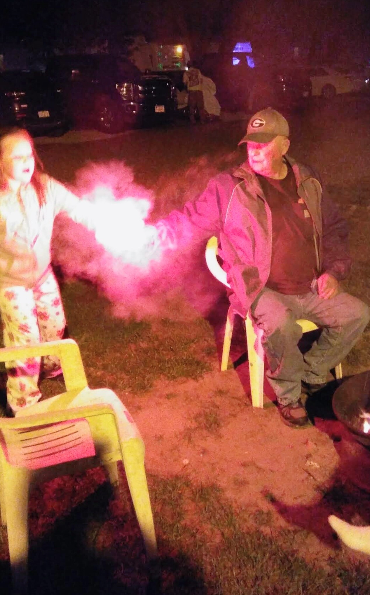 Two people outside at night; a man in a green cap and jacket sits on a chair while a pink smoke flare emits a pink cloud nearby, with a woman on the left.