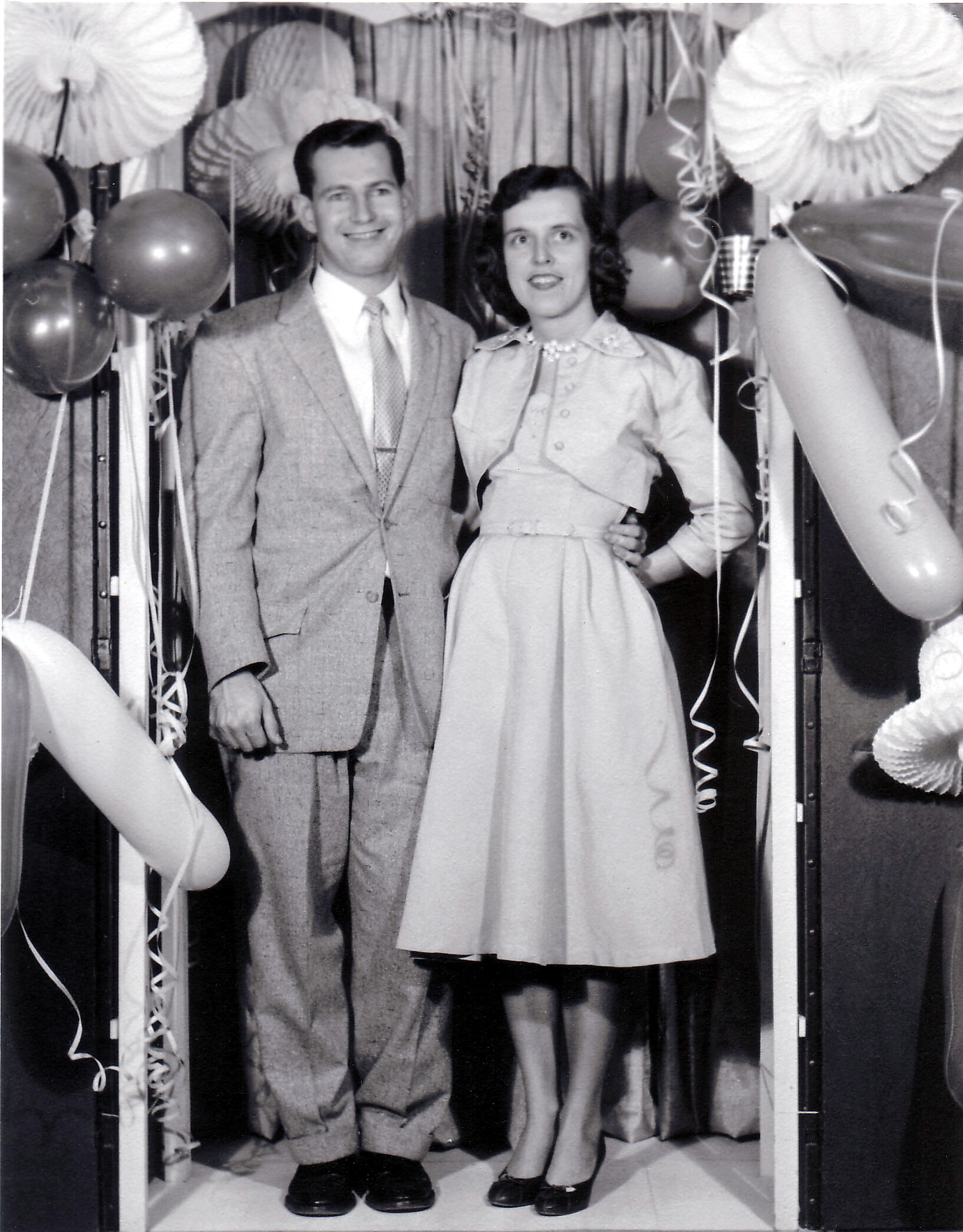 Couple posing at a celebration, man in a suit and woman in a dress, with balloons and streamers around themor.