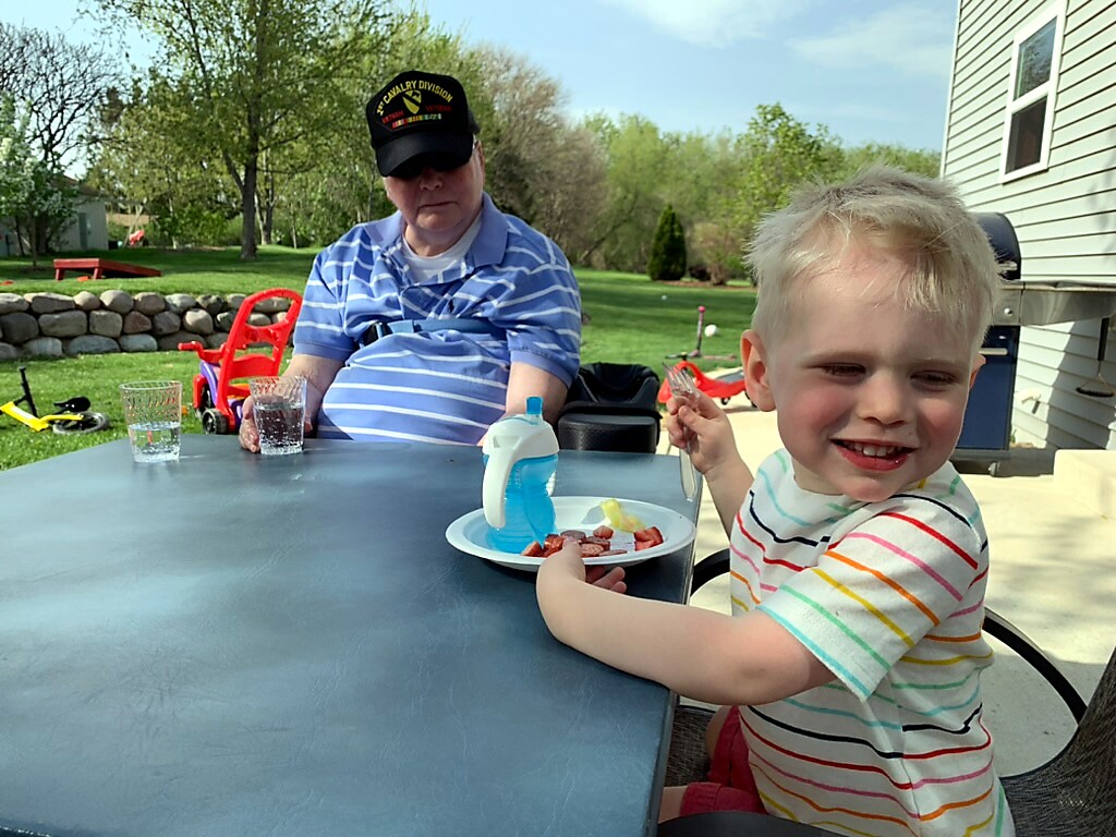 Smiling young boy in a striped shirt at a patio table outside, holding a fork over a plate of strawberries and a blue bottle; a man in a cap sits nearby in the yard.