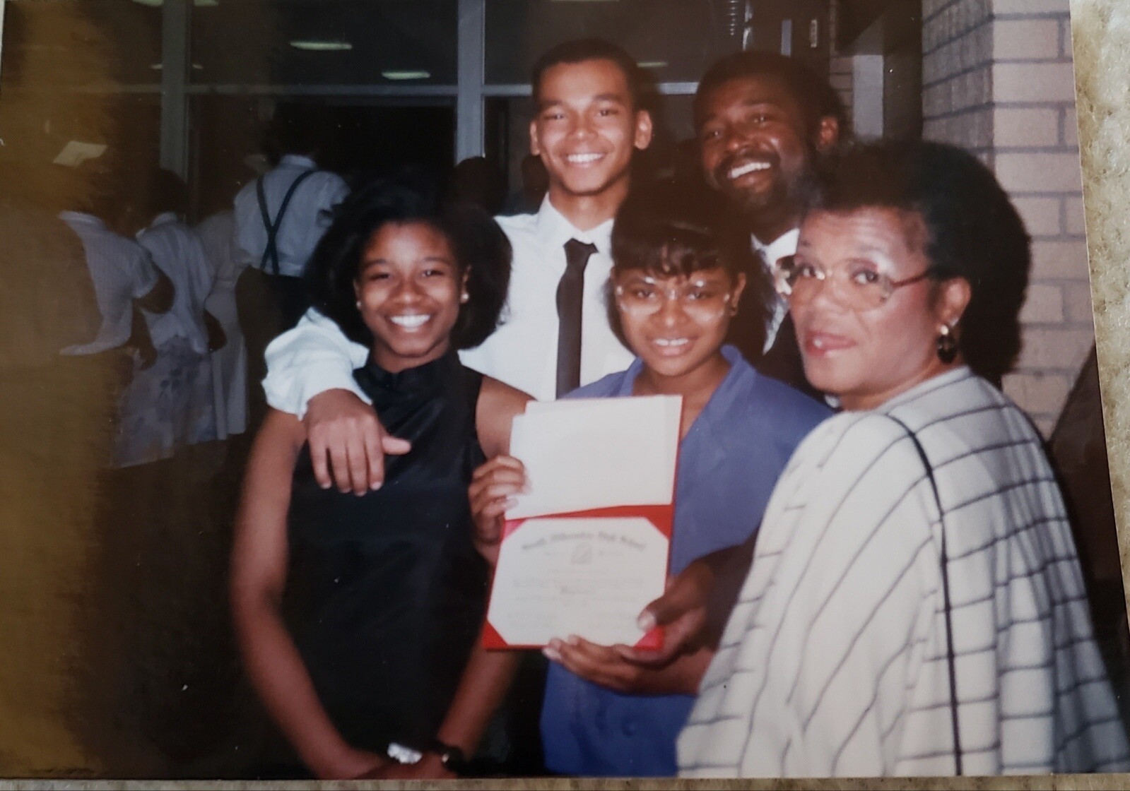Five people pose indoors for a graduation photo: two men in back, three women in front, one holding a red folder with a diploma, all smiling.