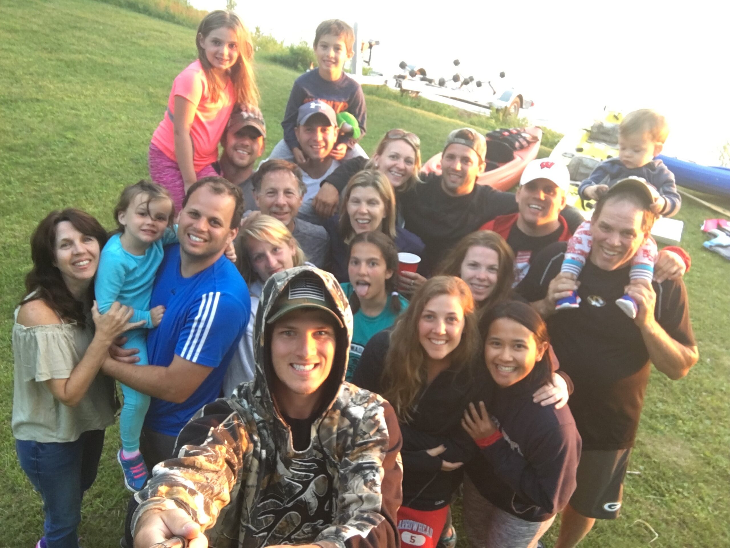 Large group of smiling people posing for a selfie on a grassy field with boats in the background, outdoors.
