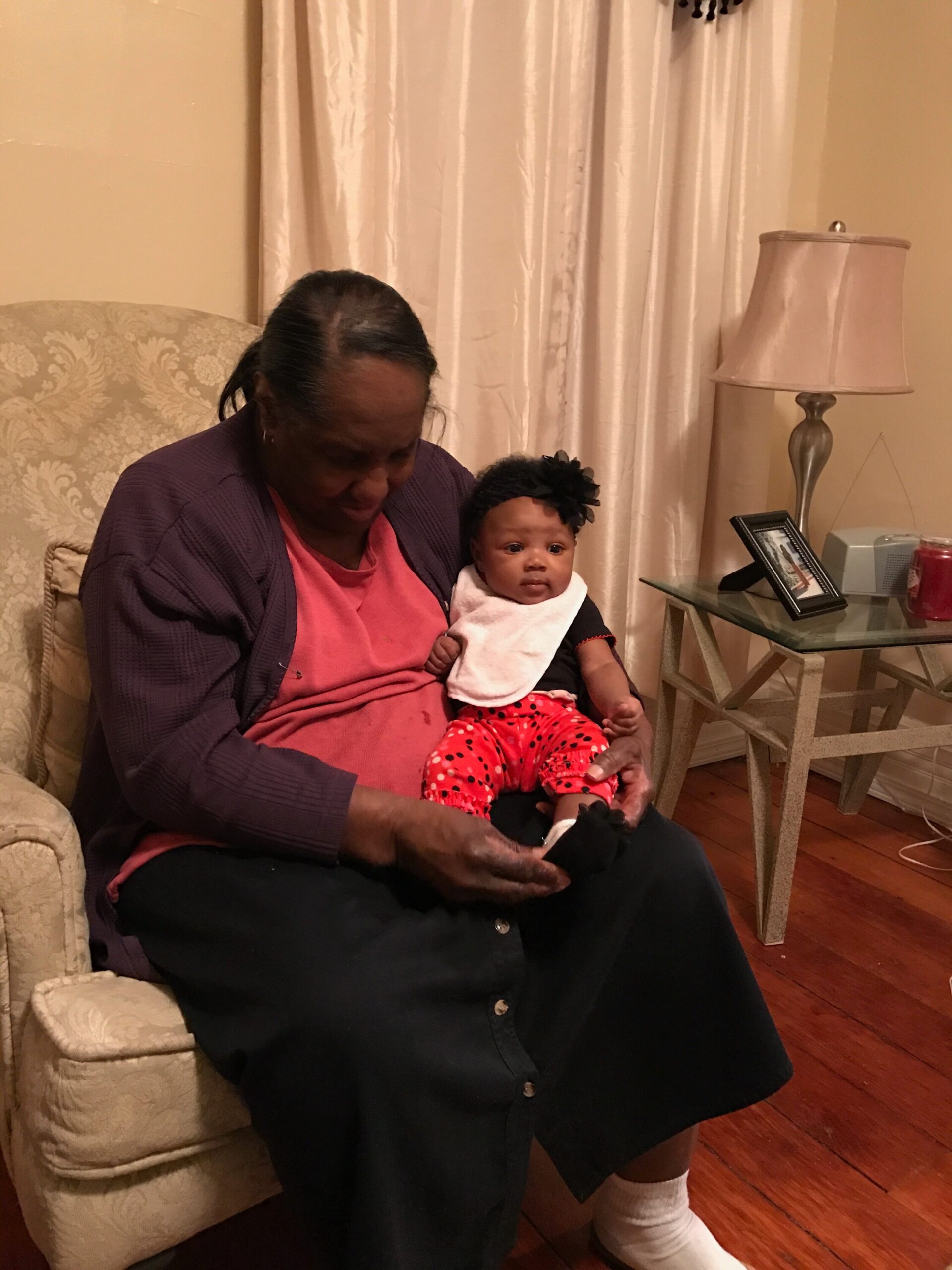 Grandmother seated in a patterned beige chair holds a baby dressed in red polka-dot pants and a white bib in a cozy living room.
