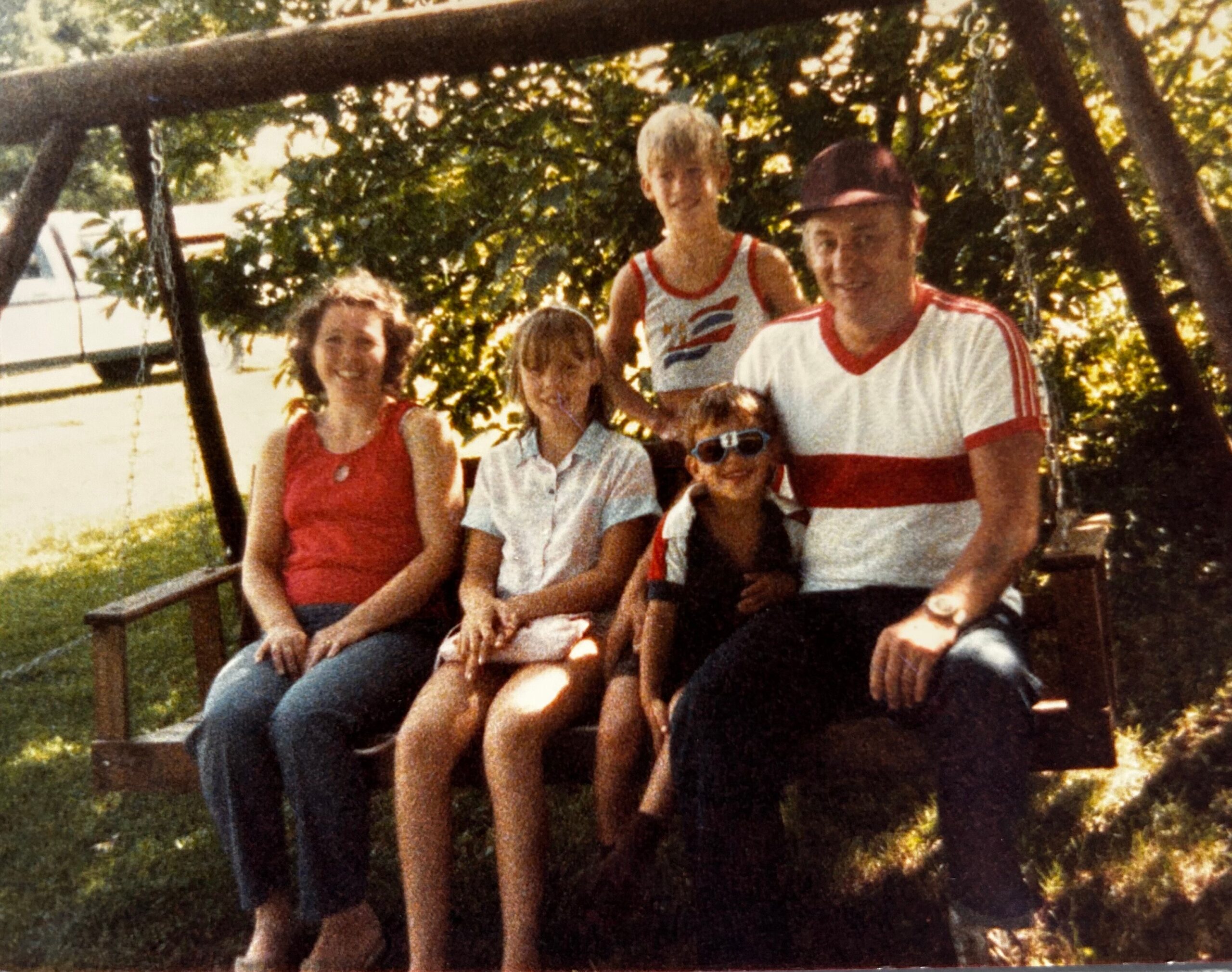 A family of six sits together on a wooden outdoor swing in a sunlit backyard, smiling at the camera.