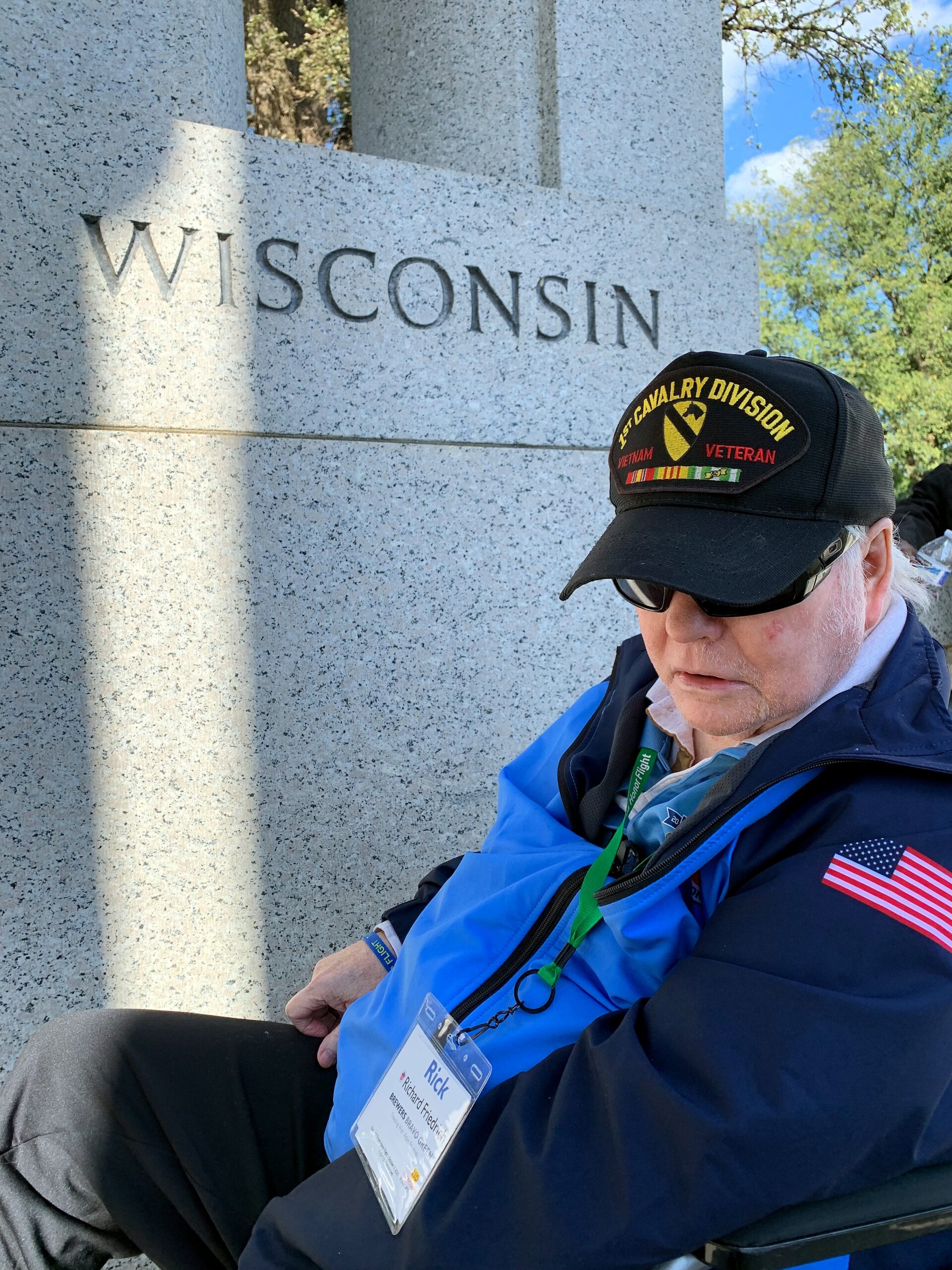 Older man in a wheelchair wearing a black cap that reads '1st Cavalry Division Vietnam Veteran' beside a granite Wisconsin monument.