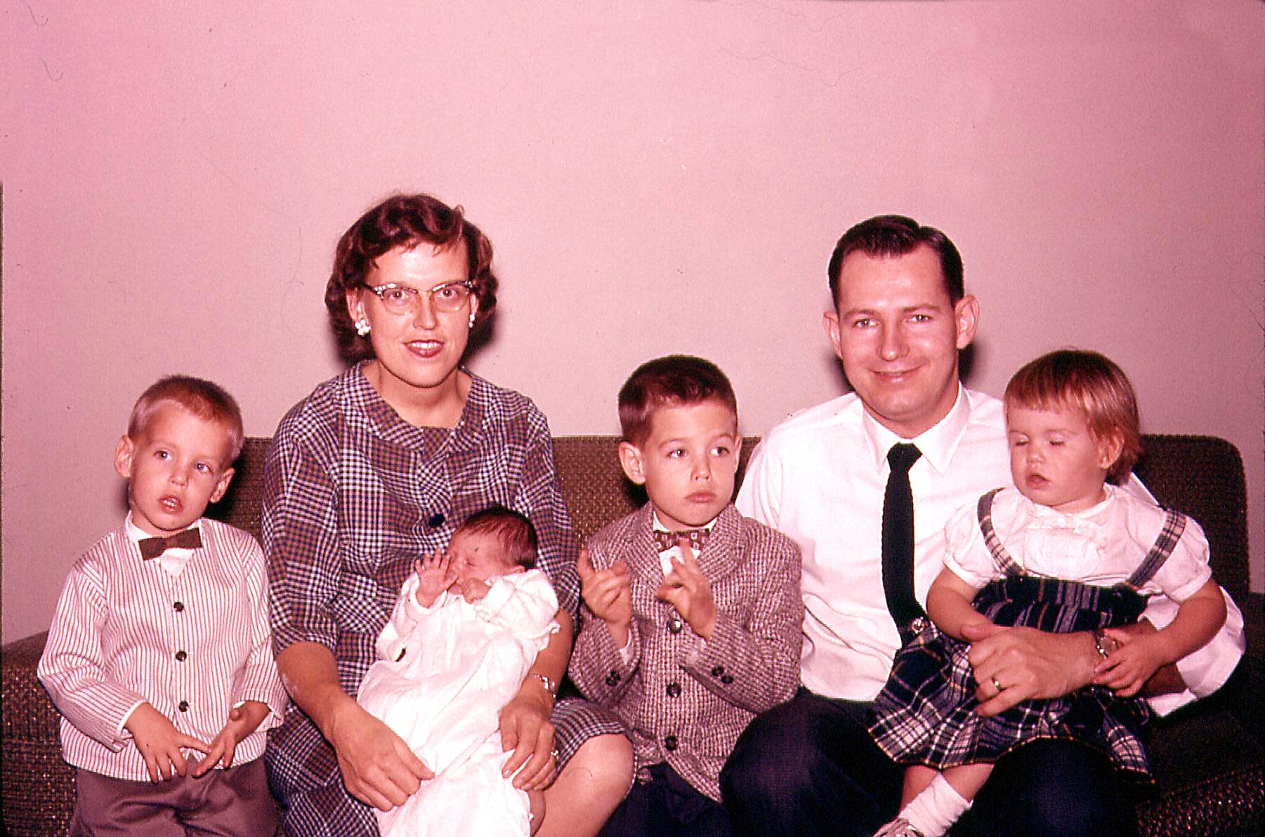 Family portrait: mother with glasses, father in a white shirt and tie, and four young children seated on a sofa indoors.