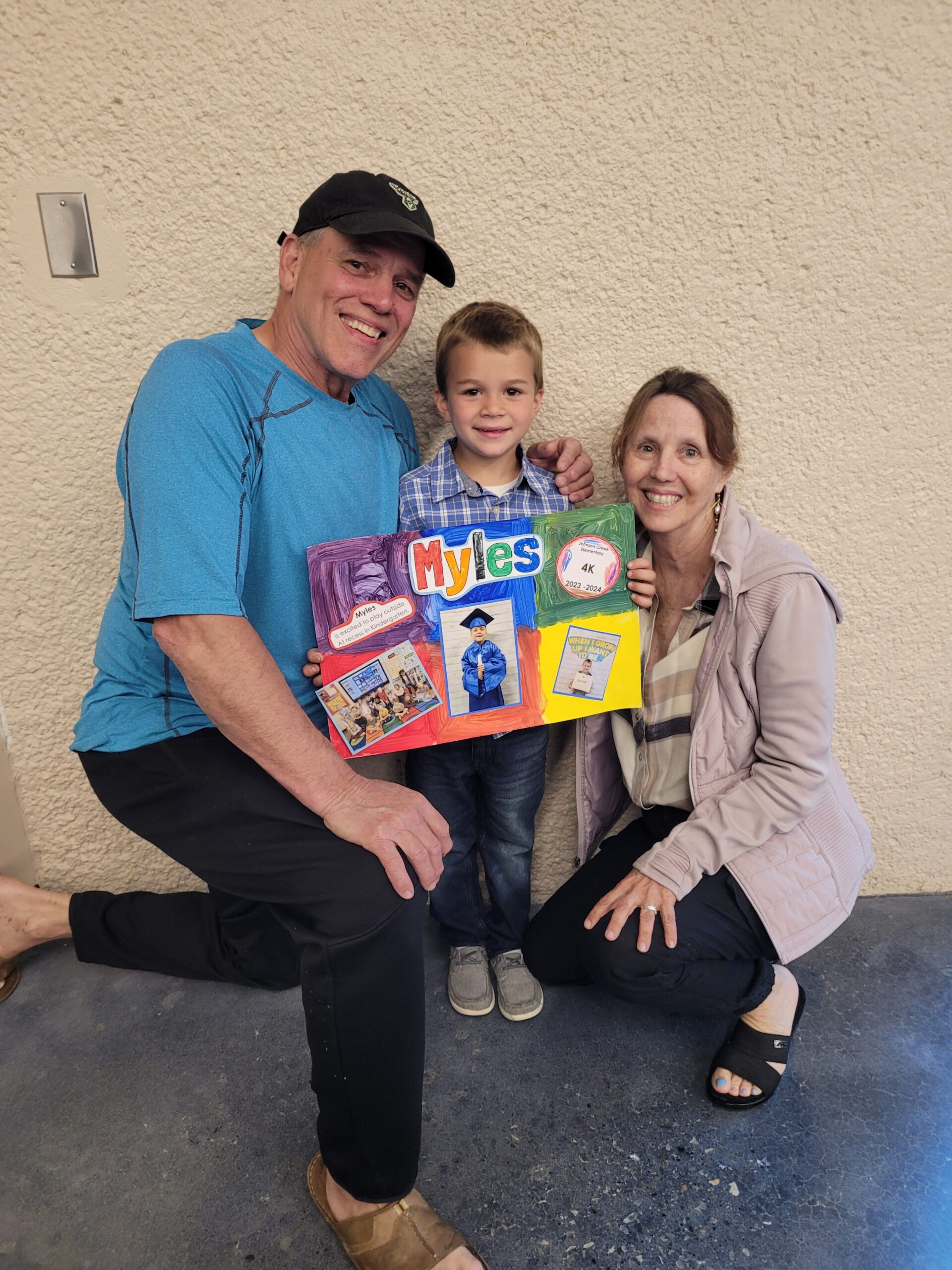 Family photo: smiling man in blue shirt and cap, a boy holding a colorful poster labeled 'Myles', and a woman kneeling beside them against a beige wall.