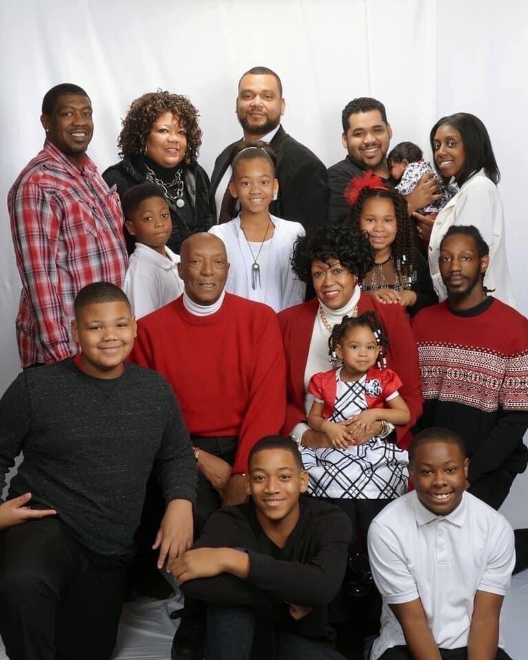 Large African American family posing for a studio group portrait in festive red, white, and black outfits, with adults and children smiling together.