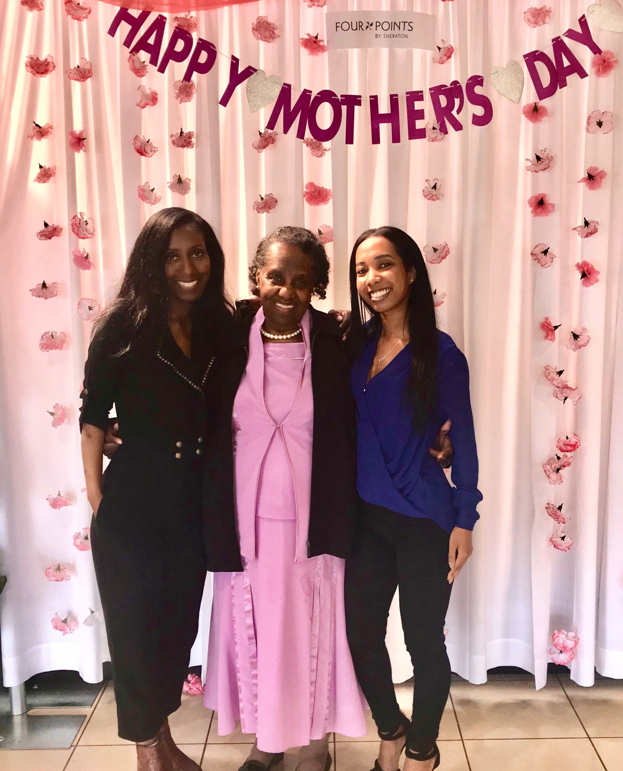 Three women pose together in front of a pink floral backdrop with a 'Happy Mother’s Day' banner, dressed in formal outfits.