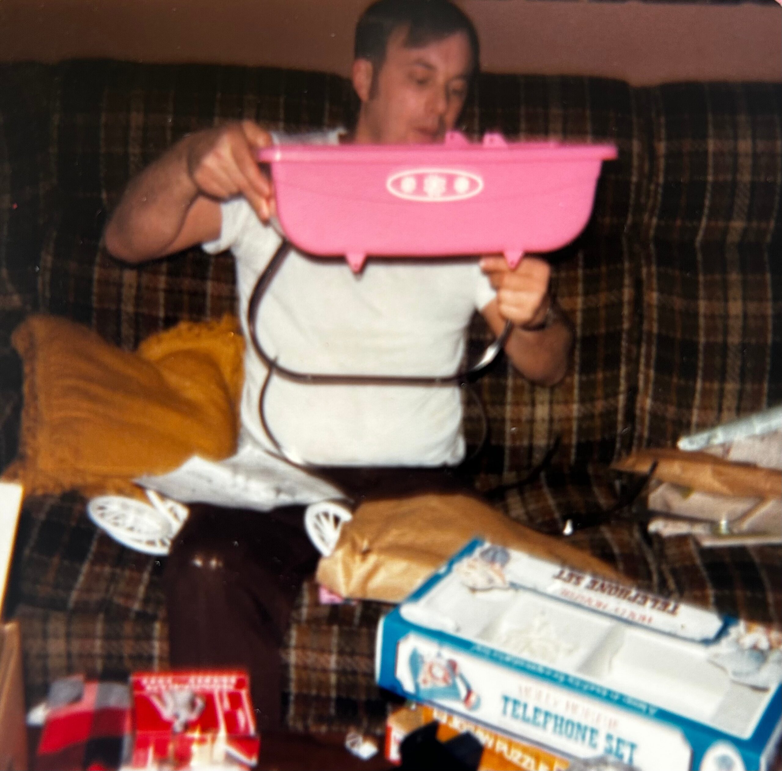 Man sitting on a plaid sofa holds a pink plastic tub with a white logo, looking down at it amid scattered items on the table/floor.