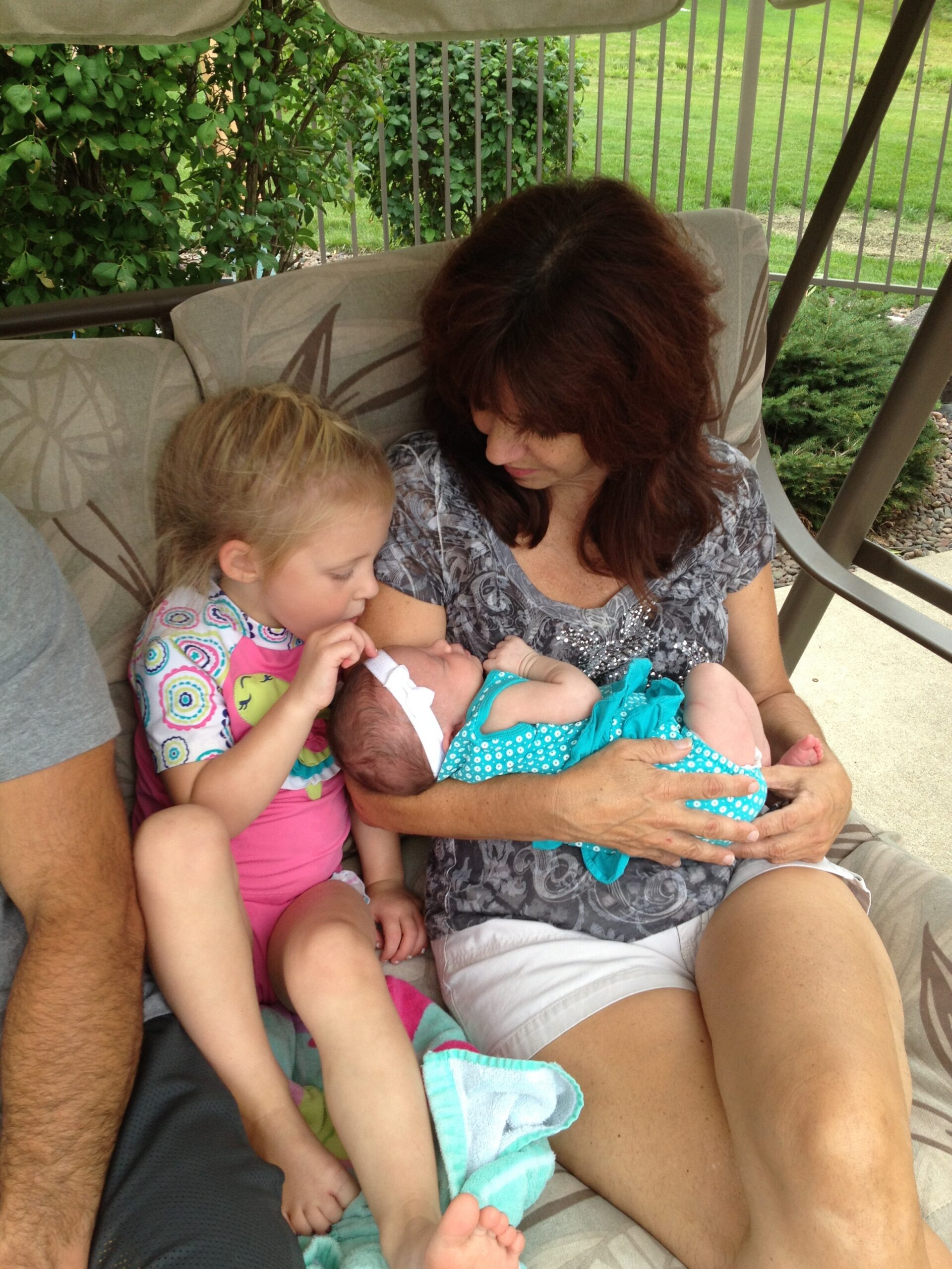 Mother on a porch swing holding a newborn baby, with a young girl beside her; greenery in the background.