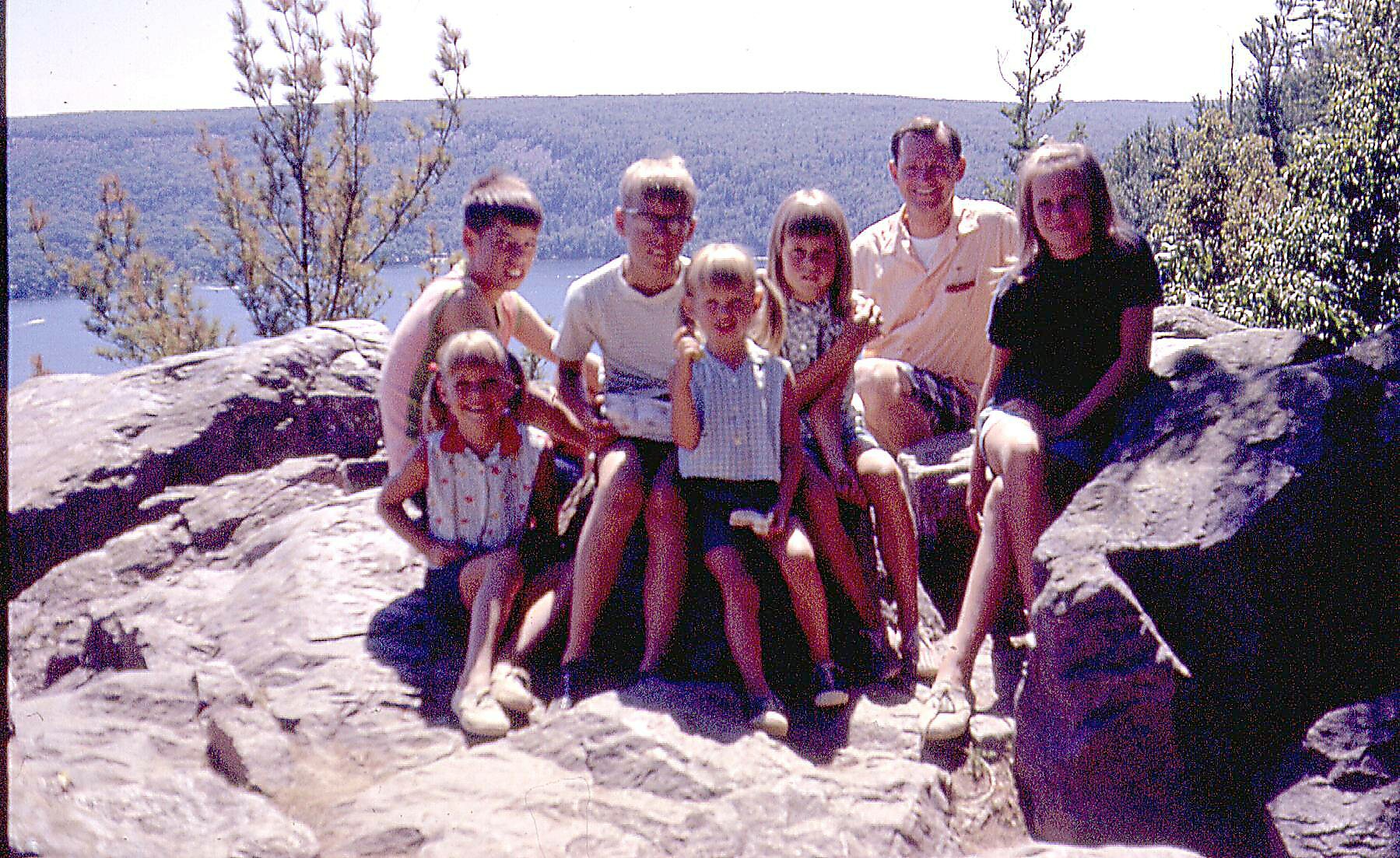 Family group portrait on a rocky overlook with a lake and forest in the background; adults and kids sit together smiling at the camera.