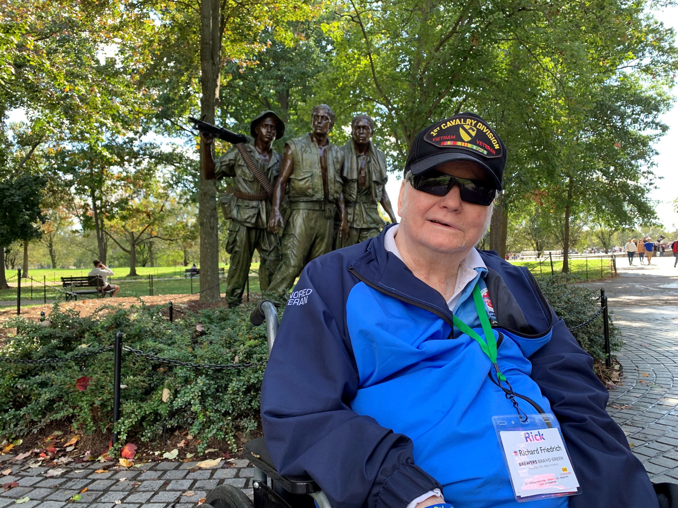 Man in a wheelchair wearing a blue jacket, taking a selfie in a park with a statue of three soldiers behind him; his cap reads '1st Cavalry Division Vietnam Veteran'./nSecond option with same scene and caption on hat: '1st Cavalry Division Vietnam Veteran' behind him in a sunny park.