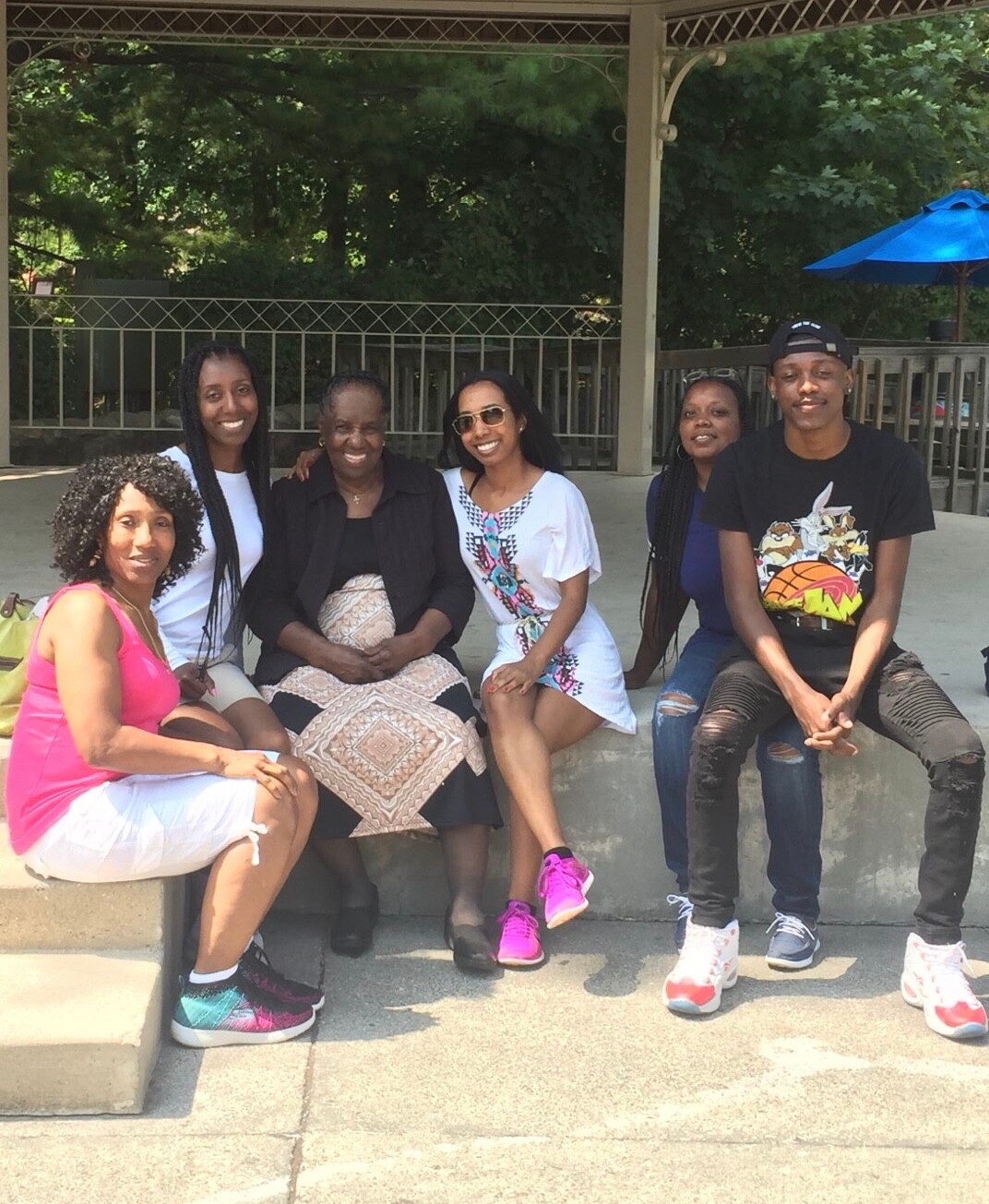 Six people sit together on a concrete ledge outdoors, smiling at the camera under a shaded pavilion.