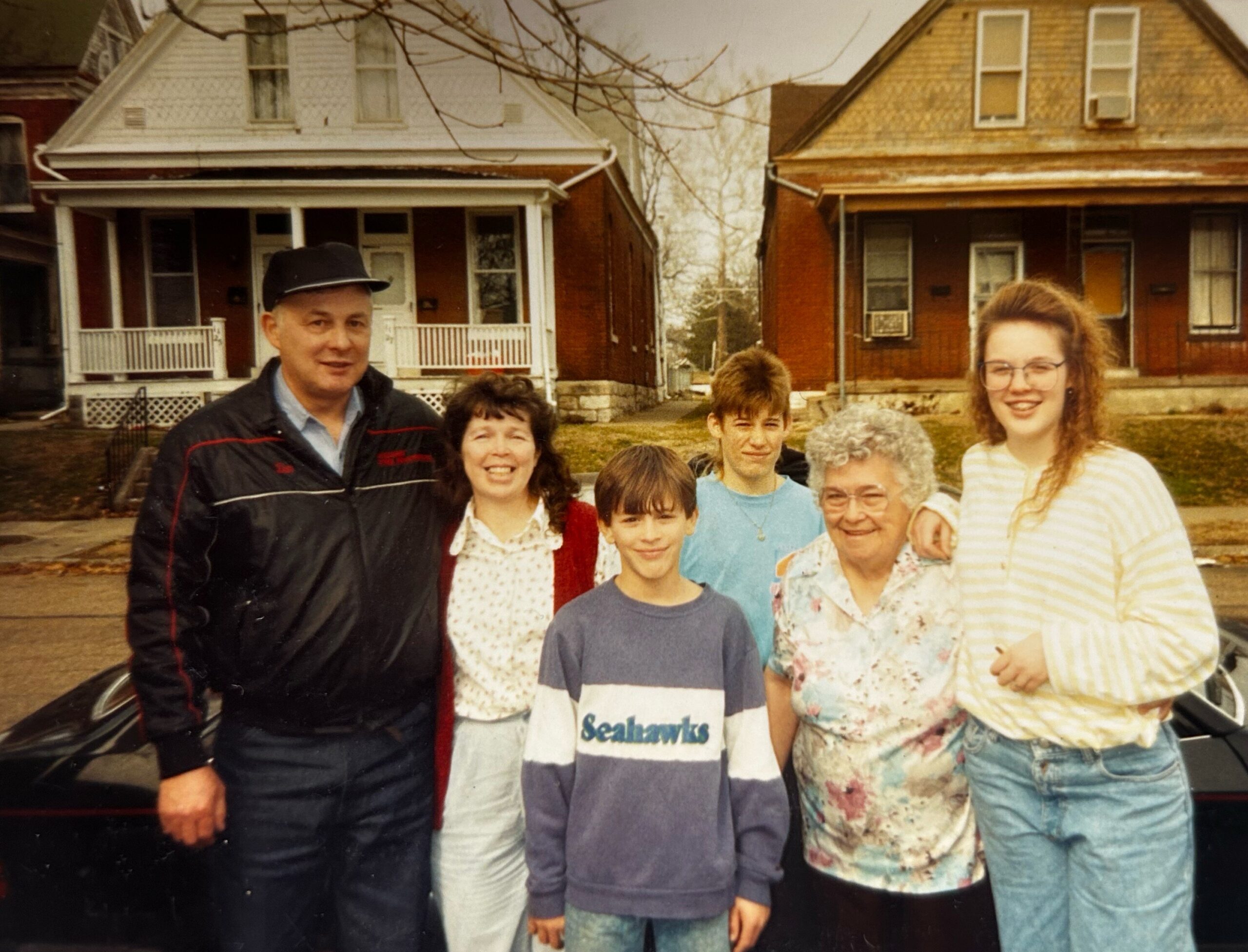 Group of seven people posing on a sidewalk in front of two brick houses, smiling at the camera.