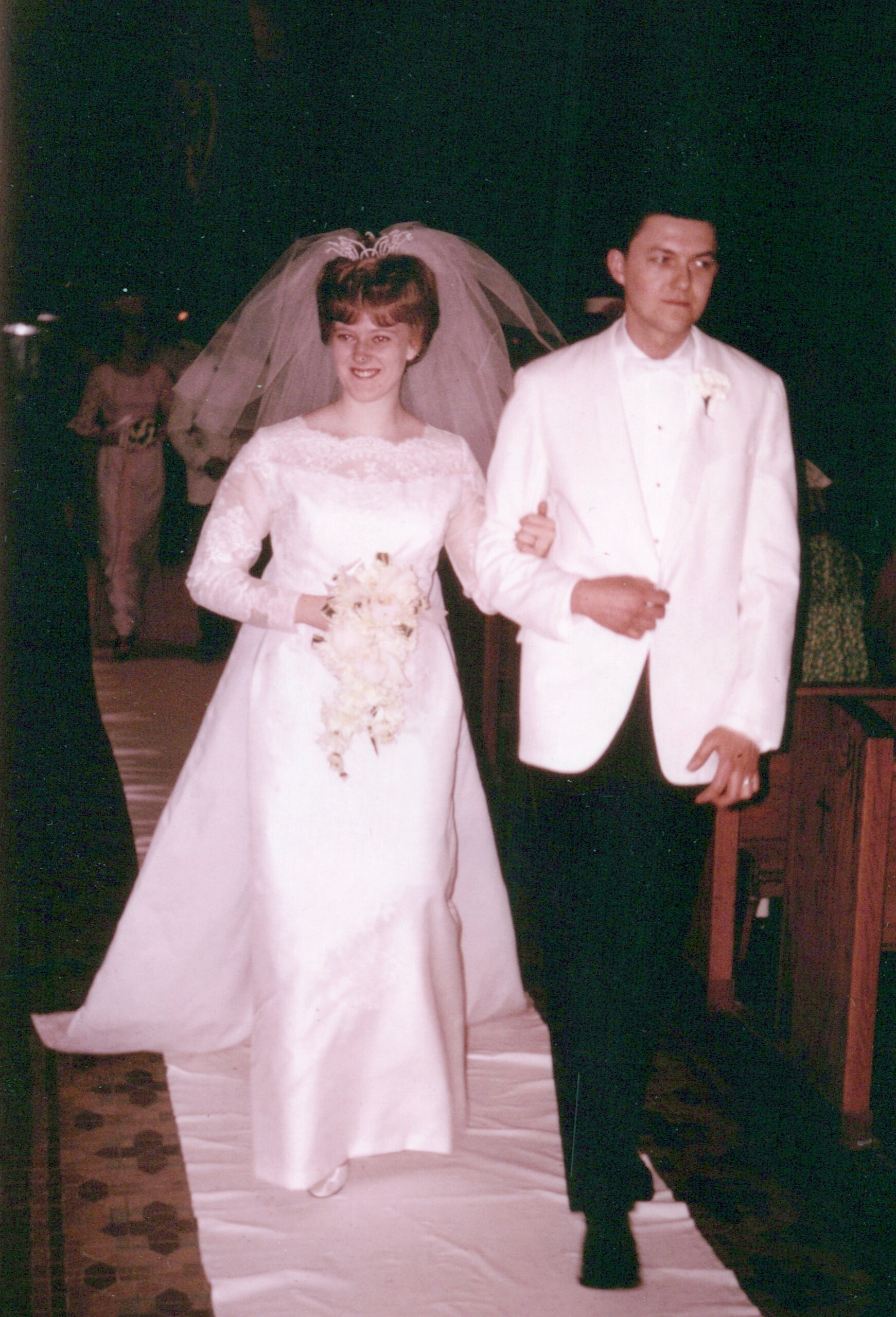 Bride in a white wedding gown and veil, holding a bouquet, walking arm-in-arm with a groom in a white tuxedo down an aisle at night.