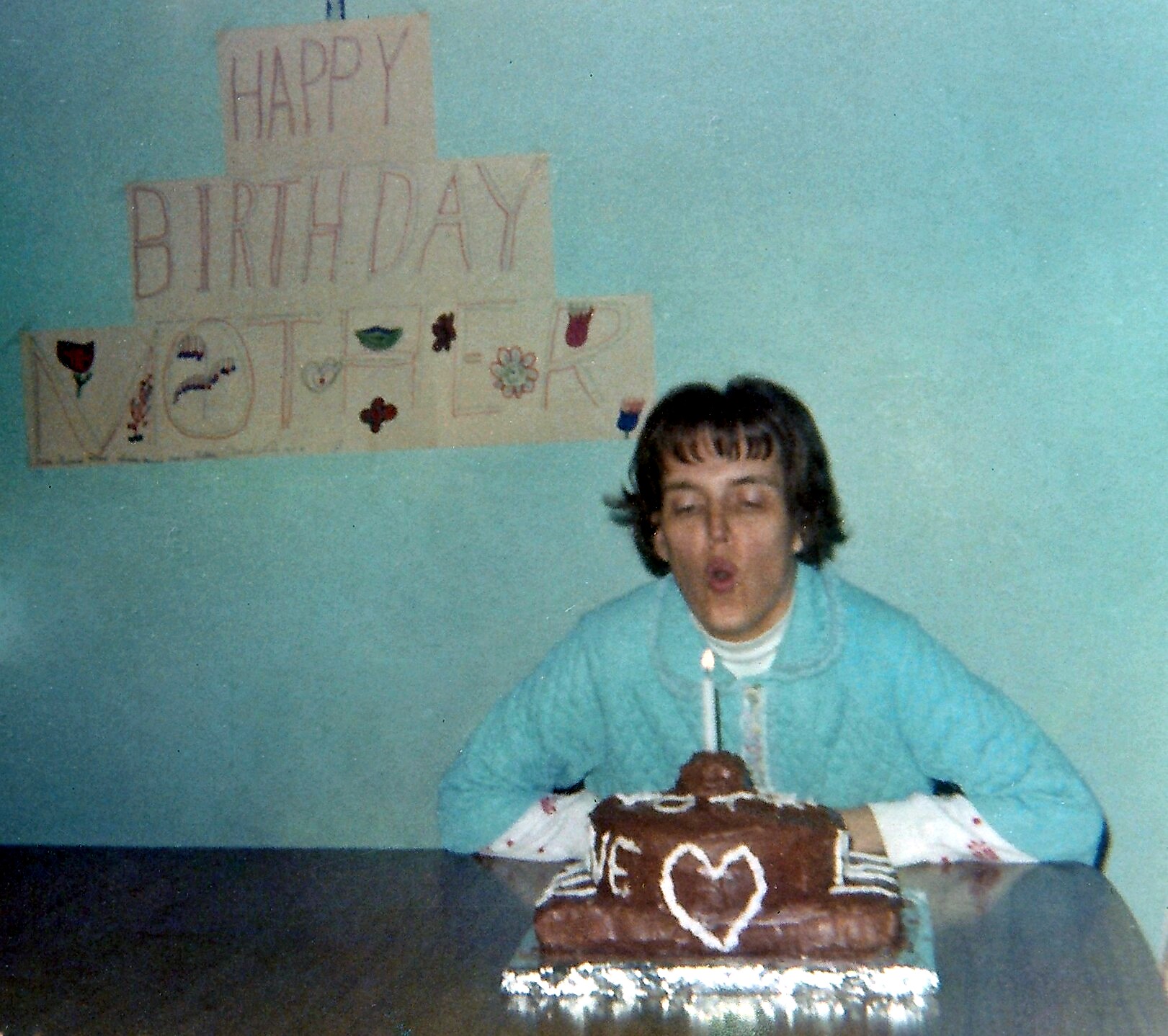 Young person in a turquoise hoodie blowing out candles on a chocolate birthday cake with a white heart decoration, at a party with a homemade 'Happy Birthday' banner on the wall behind.