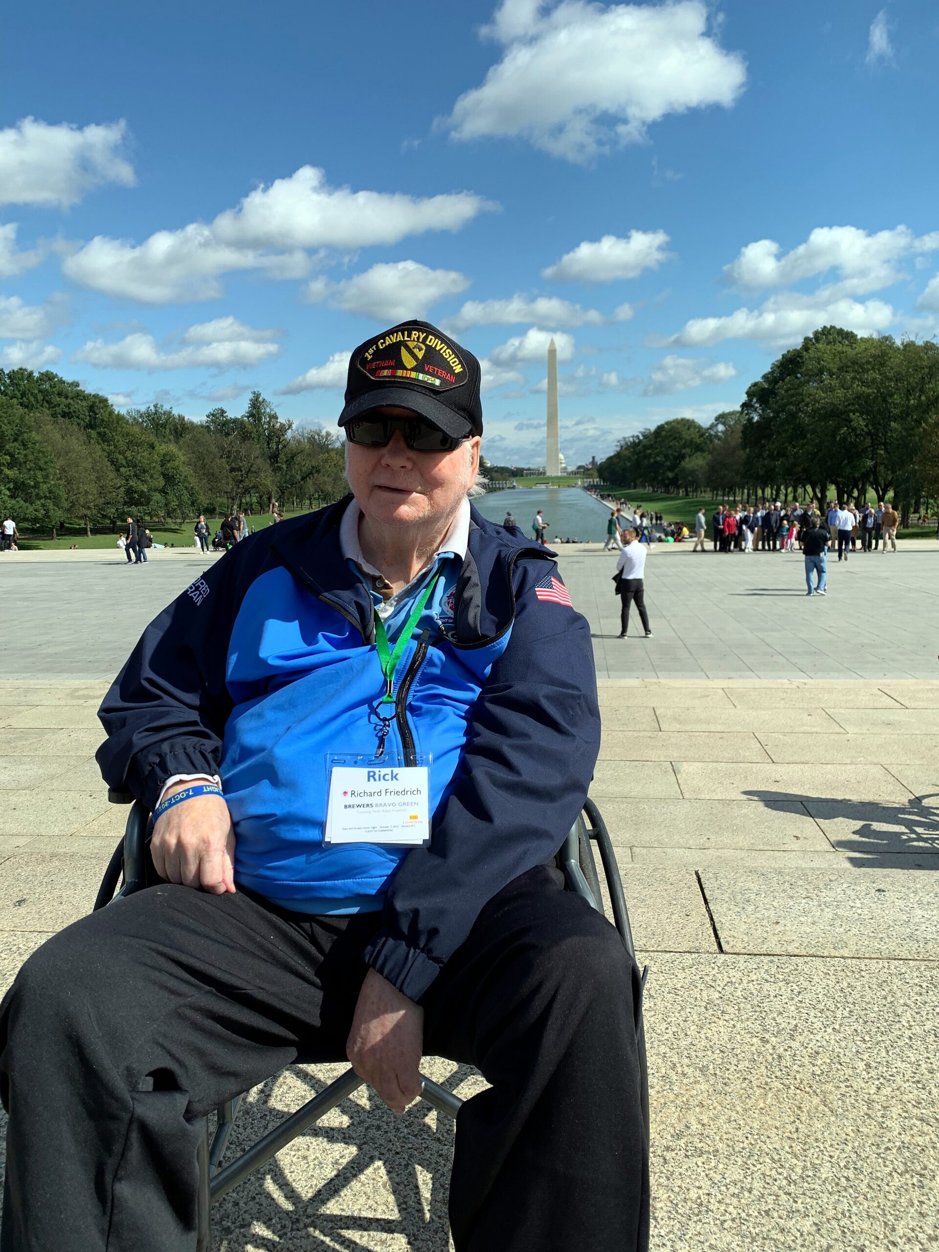 Man in a wheelchair wearing a blue jacket and a cap with a 1st Cavalry Division patch, at the National Mall with the Washington Monument in the distance under a blue sky.