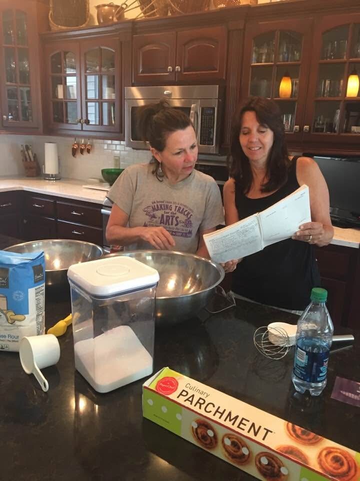 Two women in a kitchen study a recipe together, standing by large mixing bowls on a dark island with baking ingredients nearby.