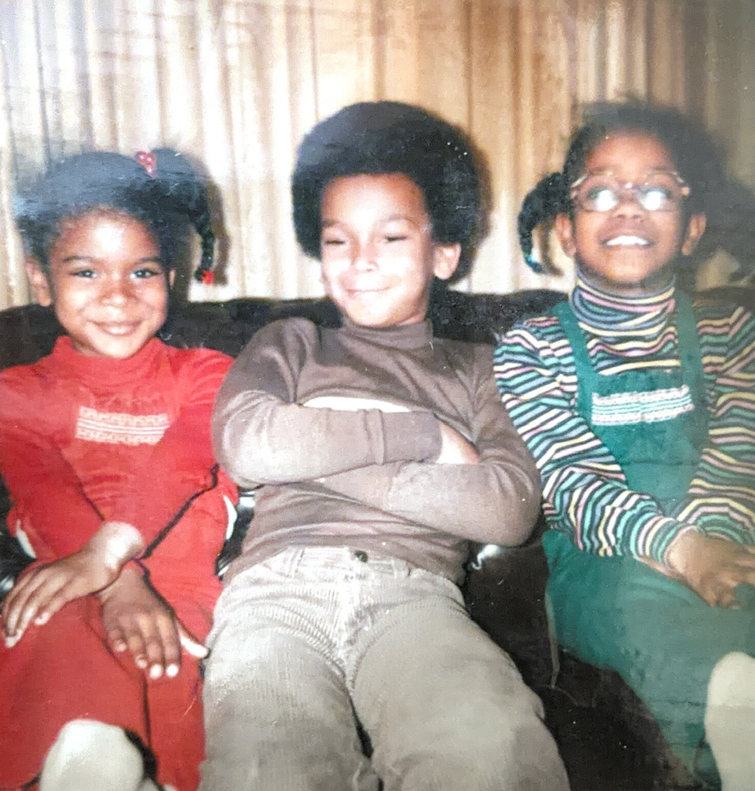 Three young children sit close together on a sofa, smiling at the camera; left in red, middle with arms crossed and Afro, right in striped shirt with green overalls and glasses.