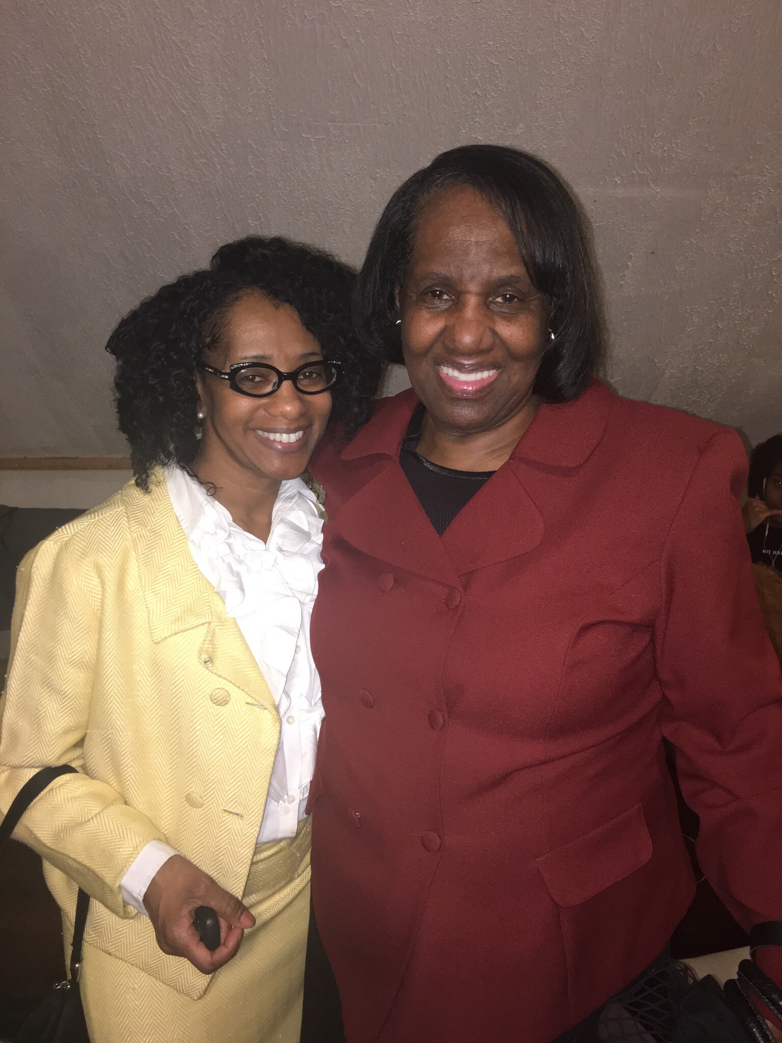 Two women posing together and smiling, one in a yellow blazer and white blouse, the other in a red suit jacket.