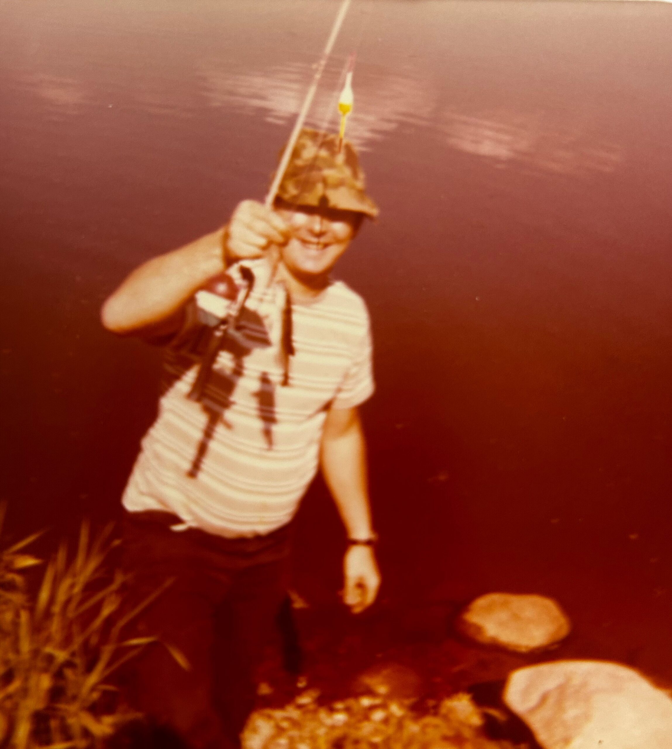 Person in a striped shirt and camouflage hat holding a fishing rod by a lake.
