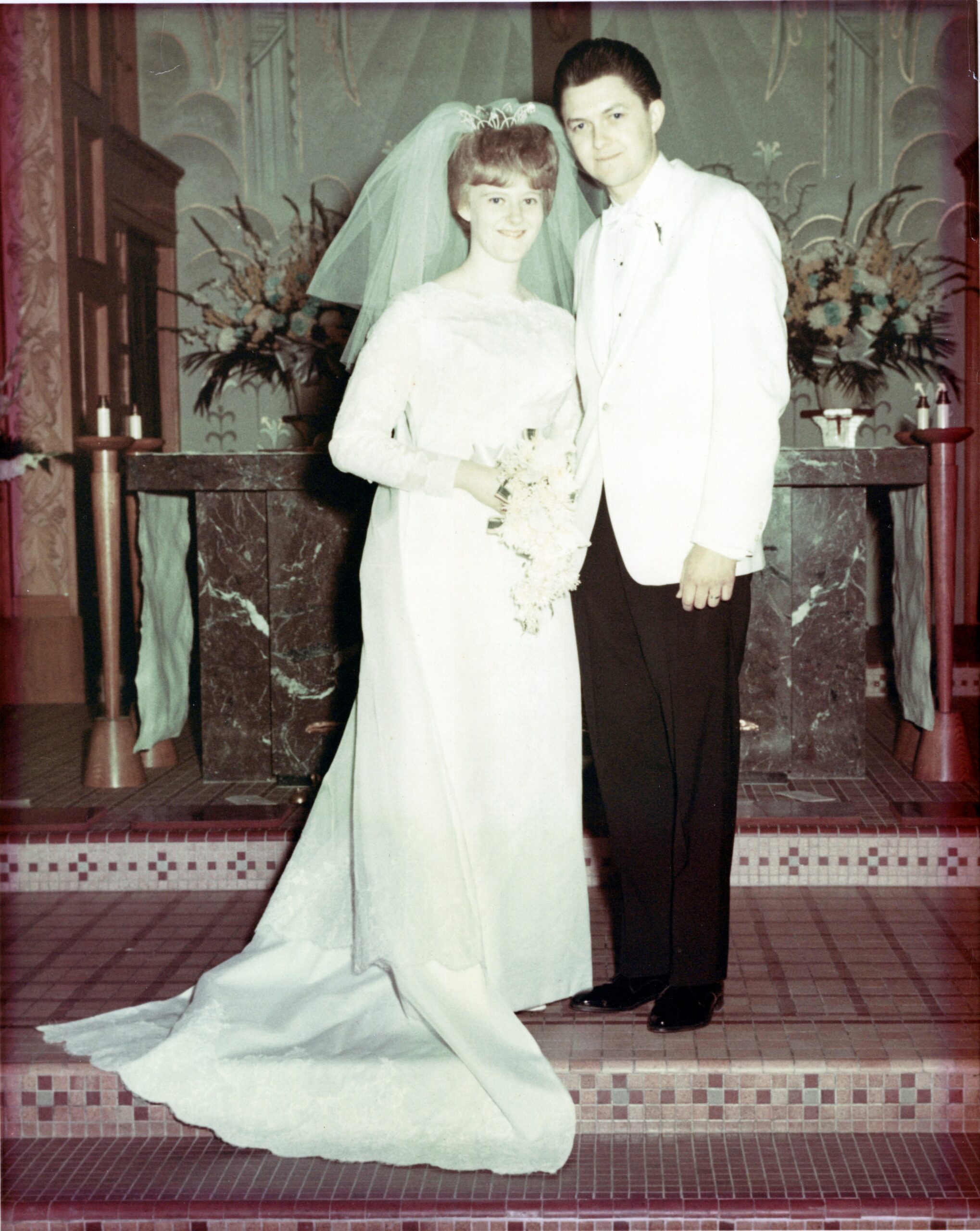 Bride in a white wedding gown with veil and bouquet stands next to a groom in a light suit on a tiled staircase at a wedding venue.