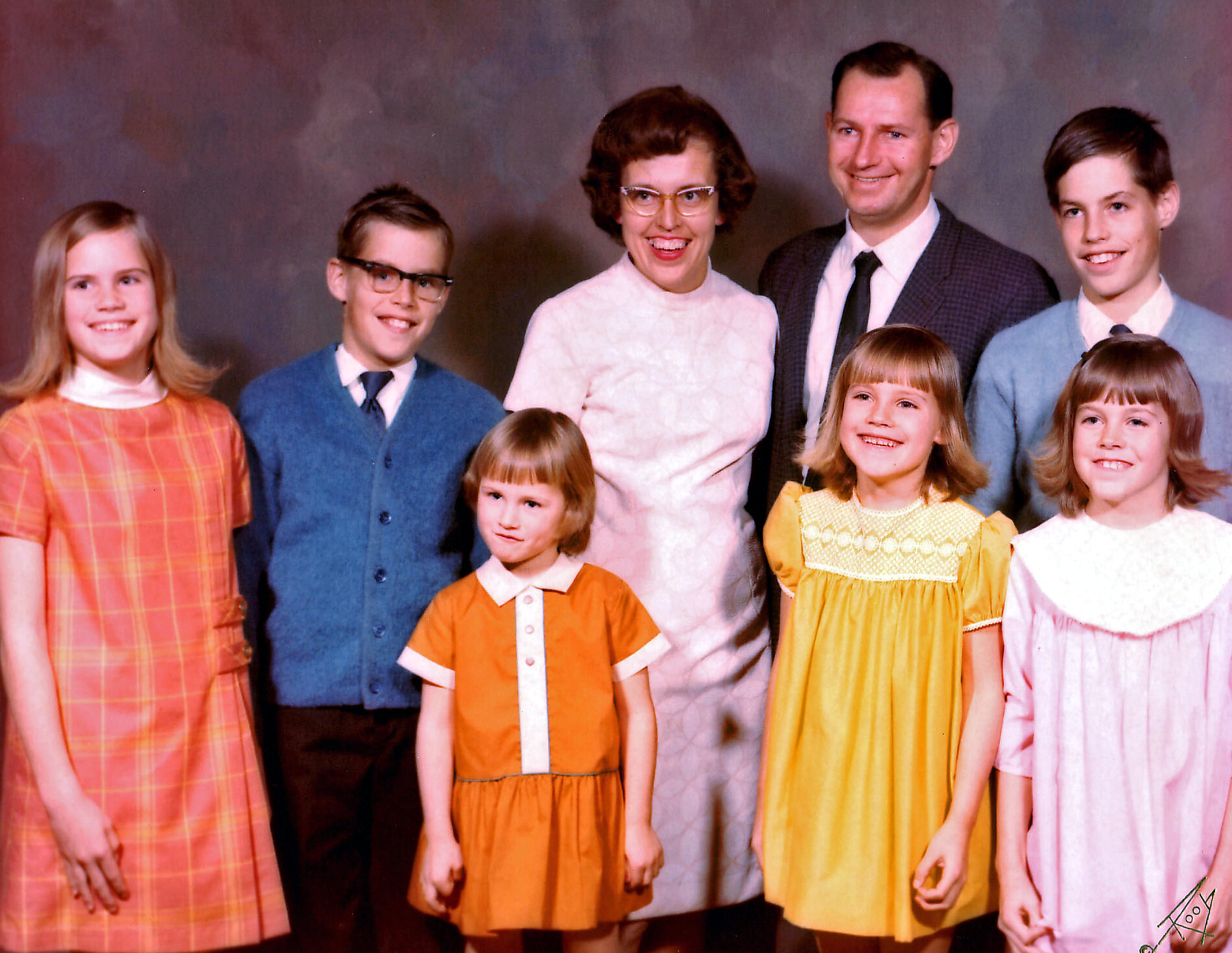 Eight people in a studio portrait: two adults and six children, all smiling and wearing 1960s-style dresses and sweaters against a neutral backdrop.