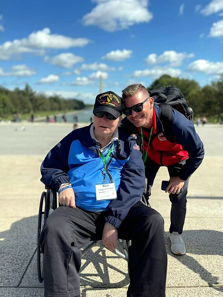Two men posing for a photo outdoors on a sunny day; one man sits in a wheelchair wearing a blue jacket and cap, the other man leans in beside him wearing a red and navy jacket and sunglasses. Both wear lanyards and smile at the camera.