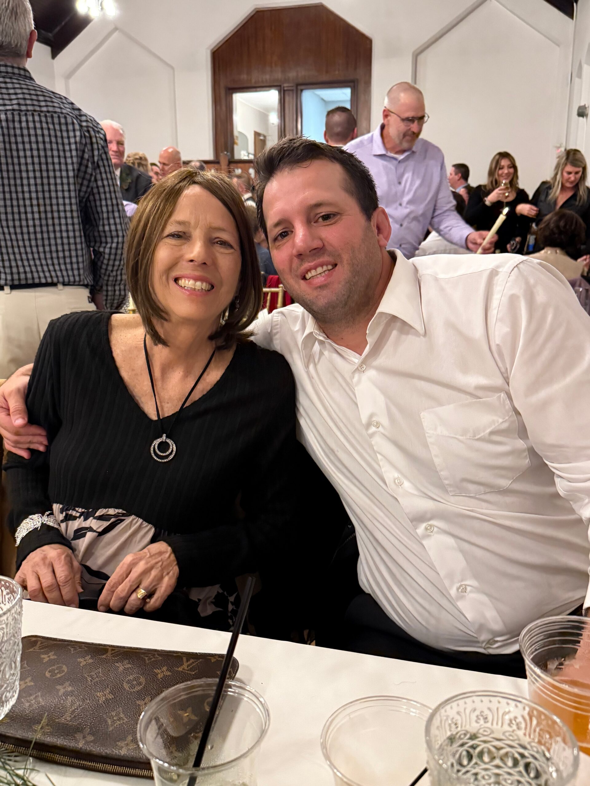 Smiling couple posing at a formal gathering, woman in a black top and necklace, man in a white shirt at a banquet table with drinks nearby.