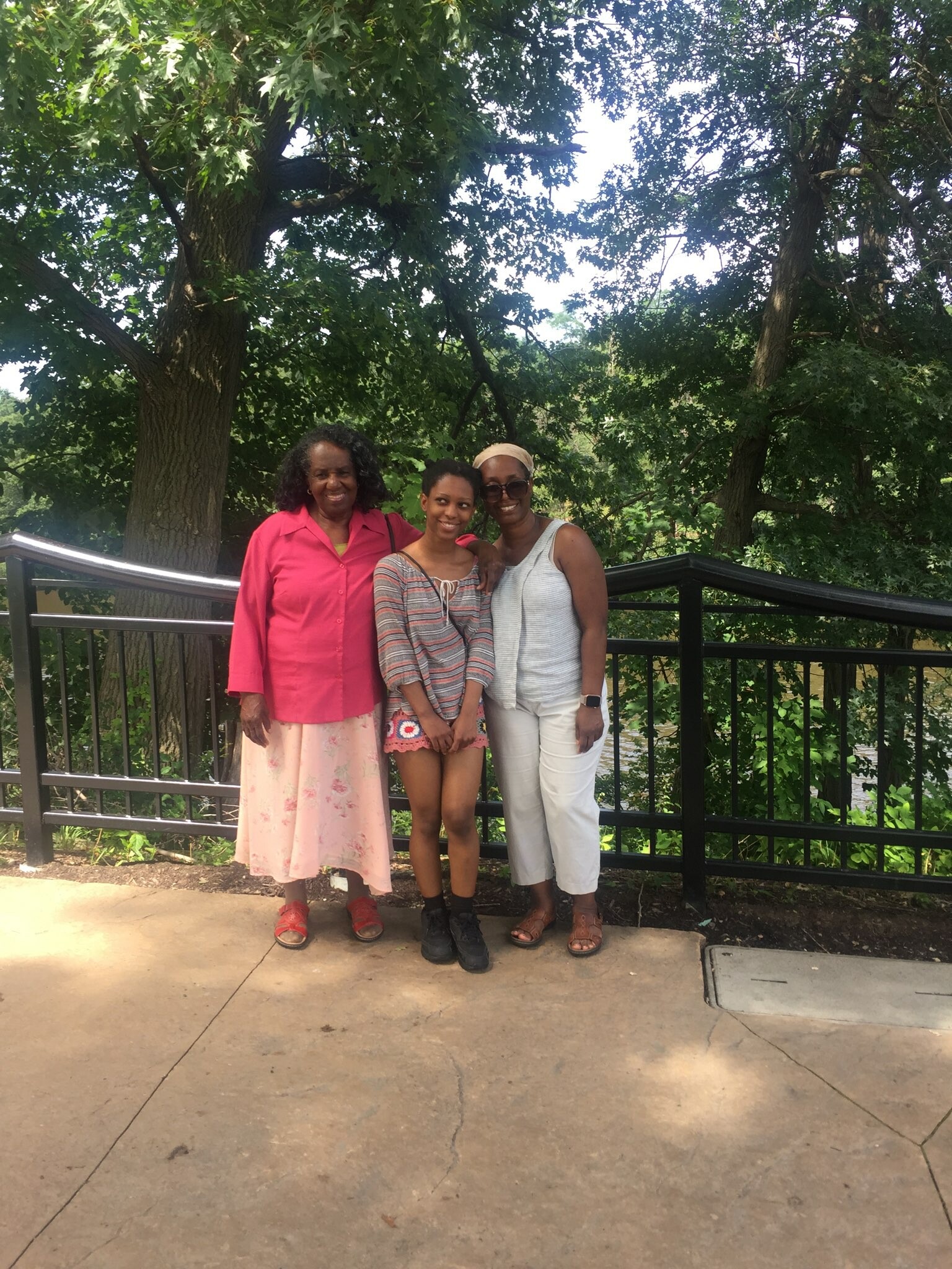 Three people stand together outdoors, smiling, in front of a black railing with green trees behind them.