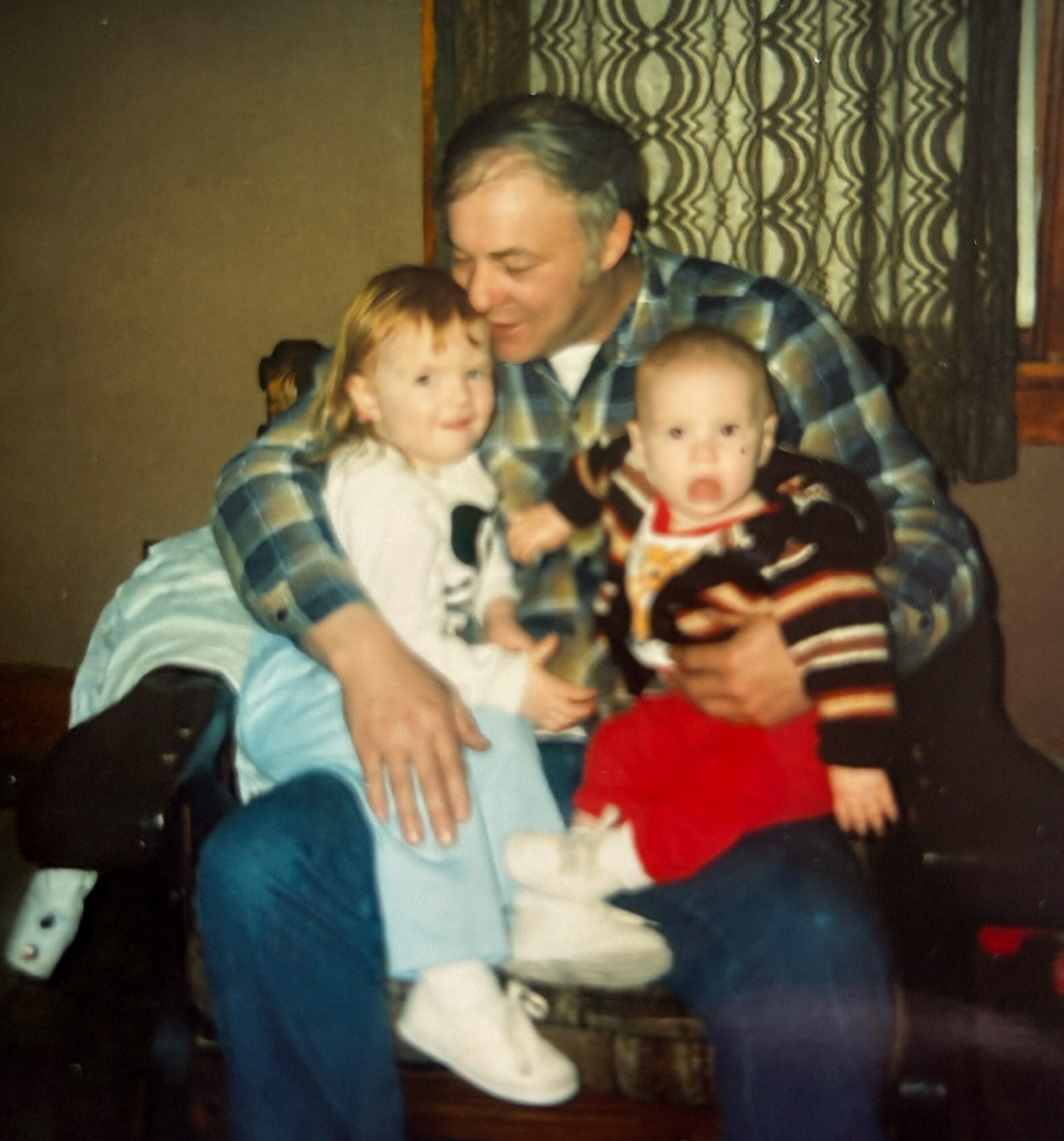 Man in plaid shirt holding two young children indoors, smiling for the camera.