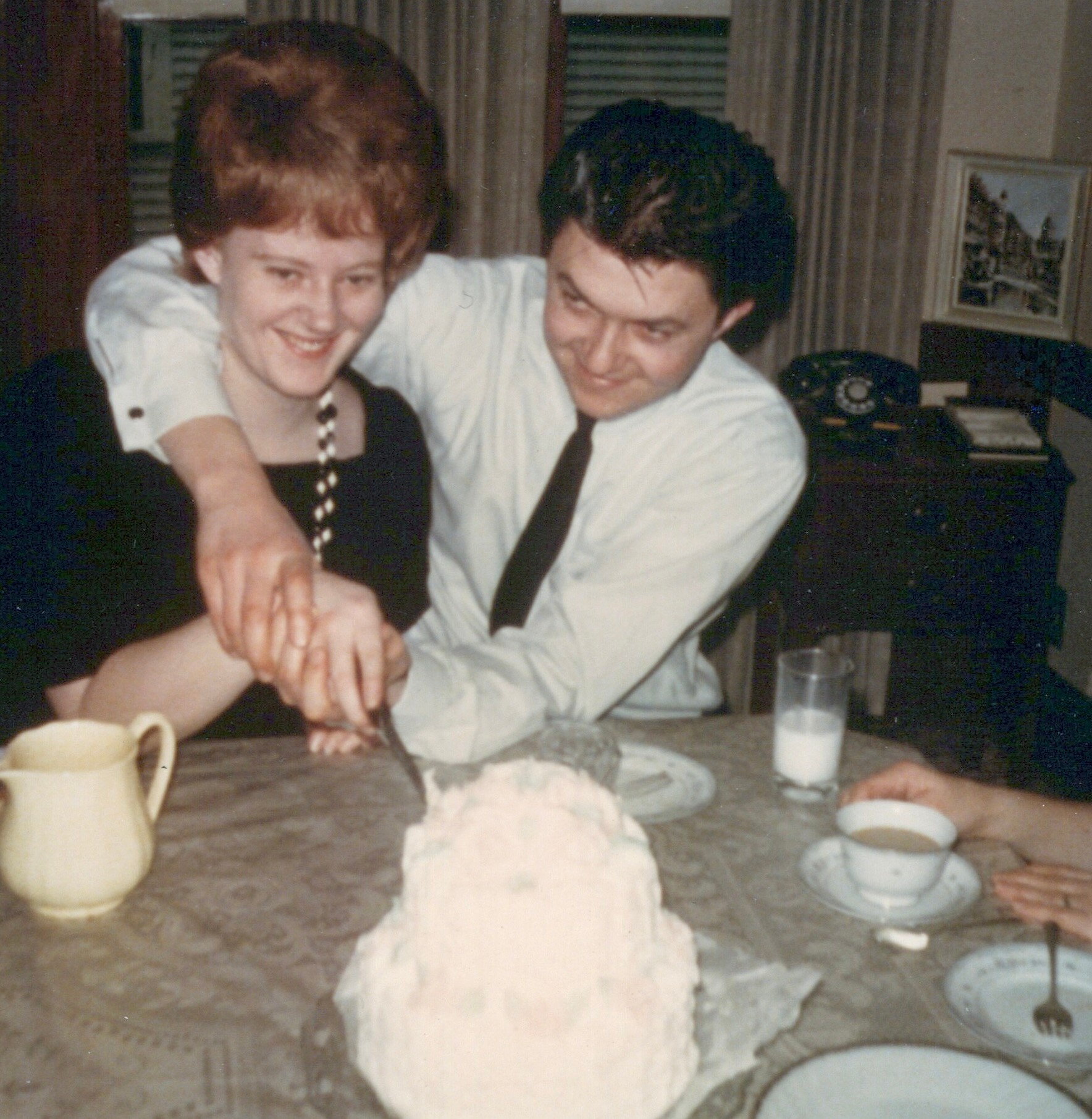 Smiling couple seated at a dining table, man with arm around woman's shoulders, cake and cups visible in the foreground.