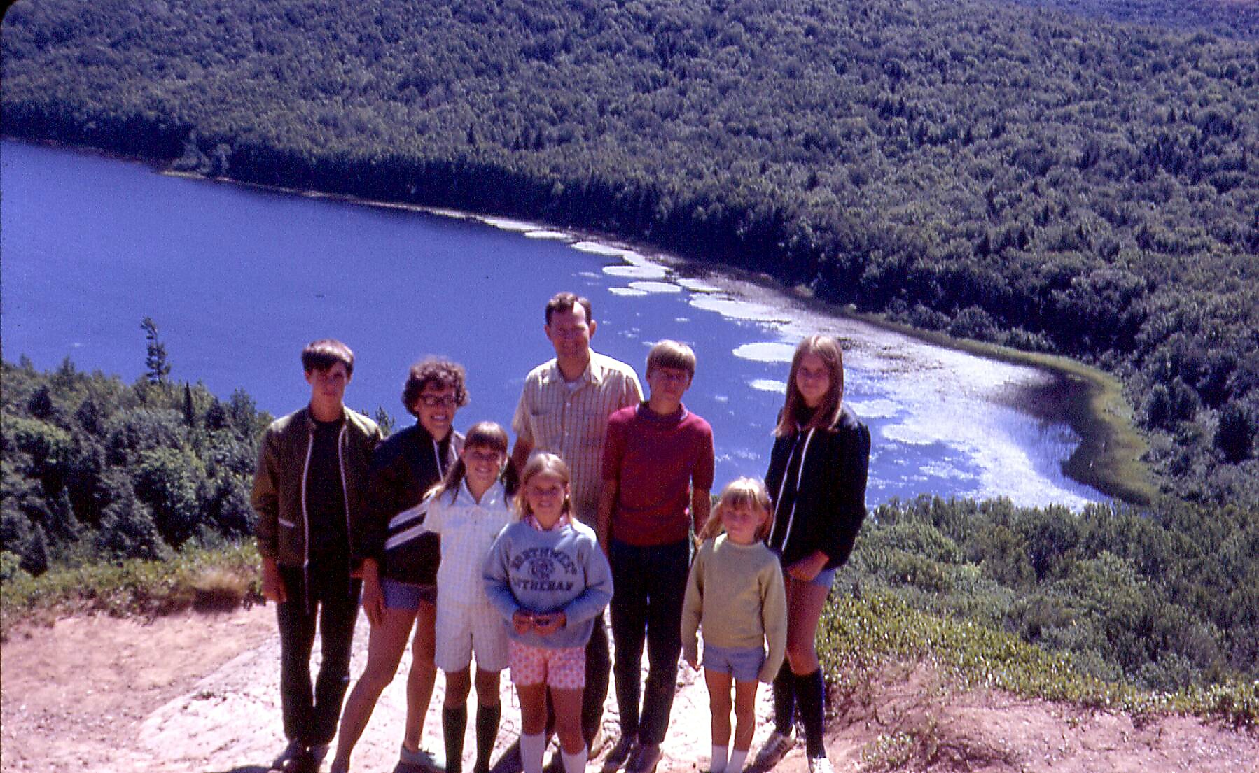 Group of nine people, kids and adults, posing on a hilltop with a lake and forest behind them.