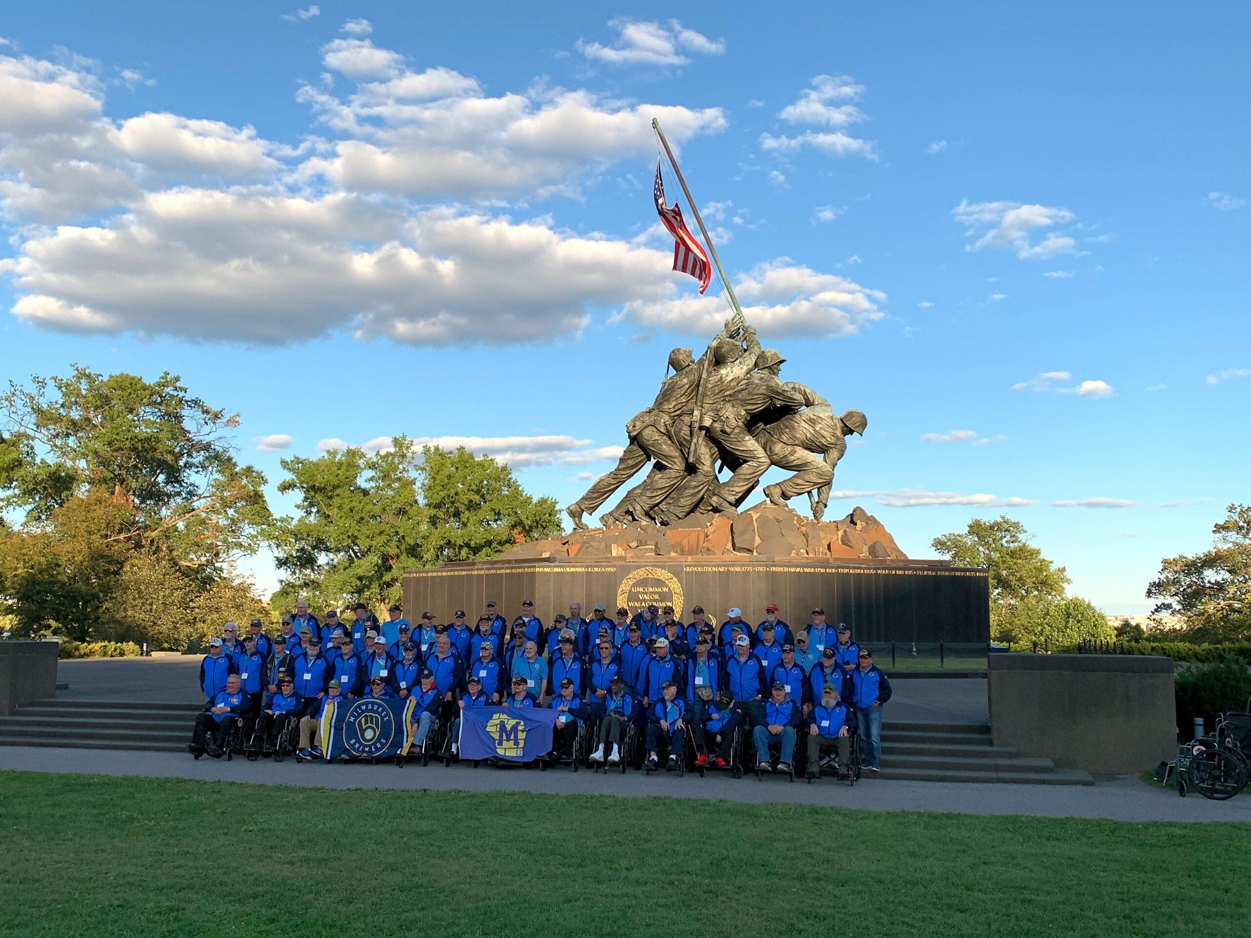 Group of people in blue jackets posing on stairs in front of a war memorial statue with an American flag raised above the figure group