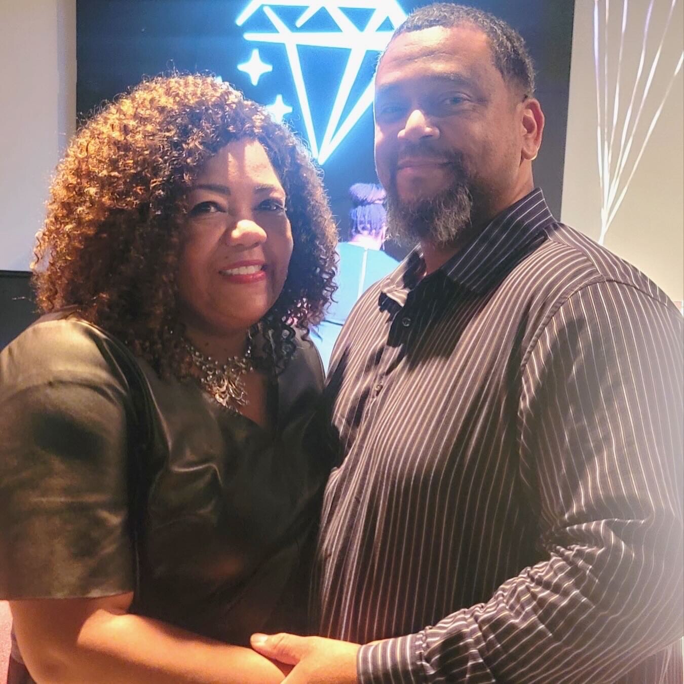Couple smiling and embracing each other indoors, woman with curly hair in a dark dress and necklace, man in a striped shirt with a short beard, neon artwork in the background