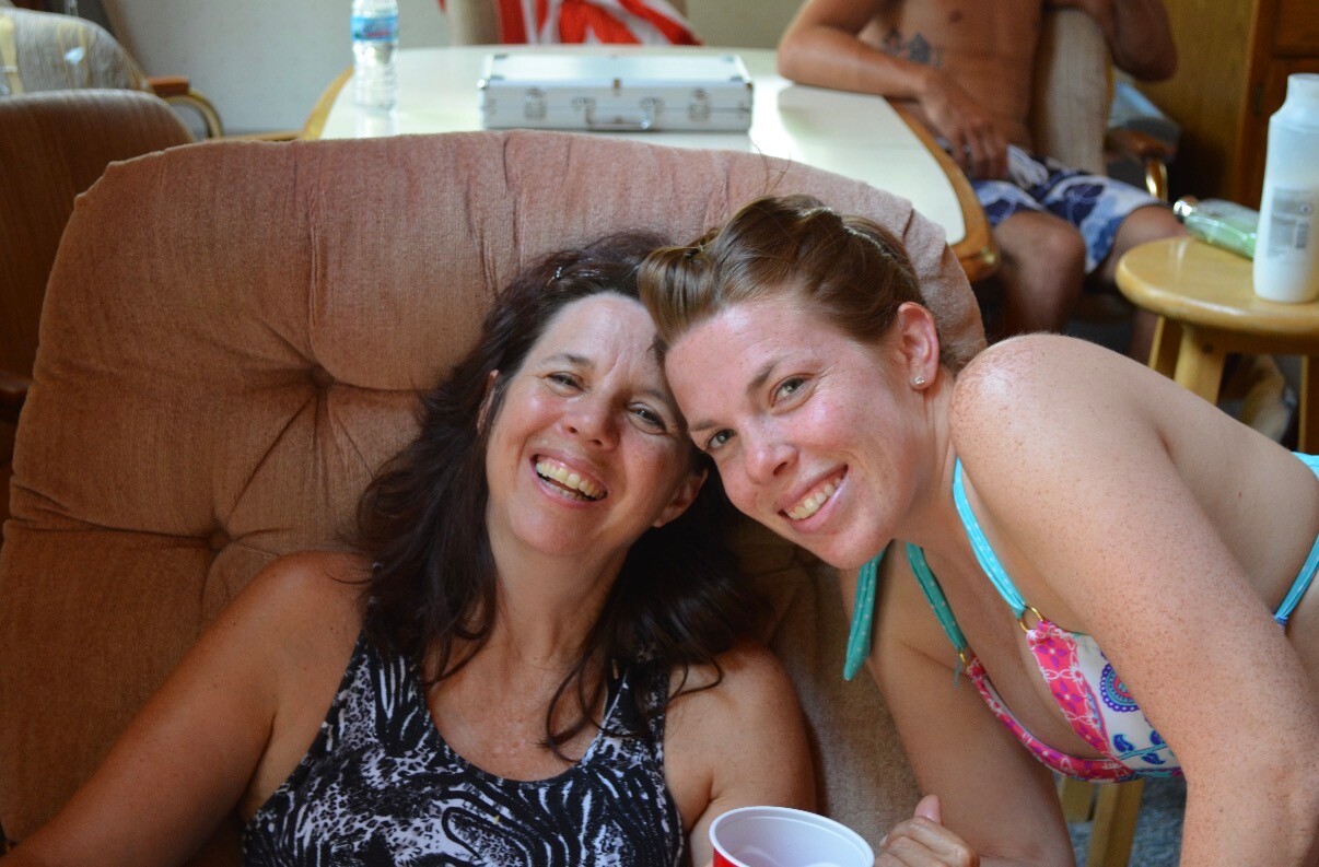 Two women in swimsuits leaning on a tan couch, smiling at the camera.