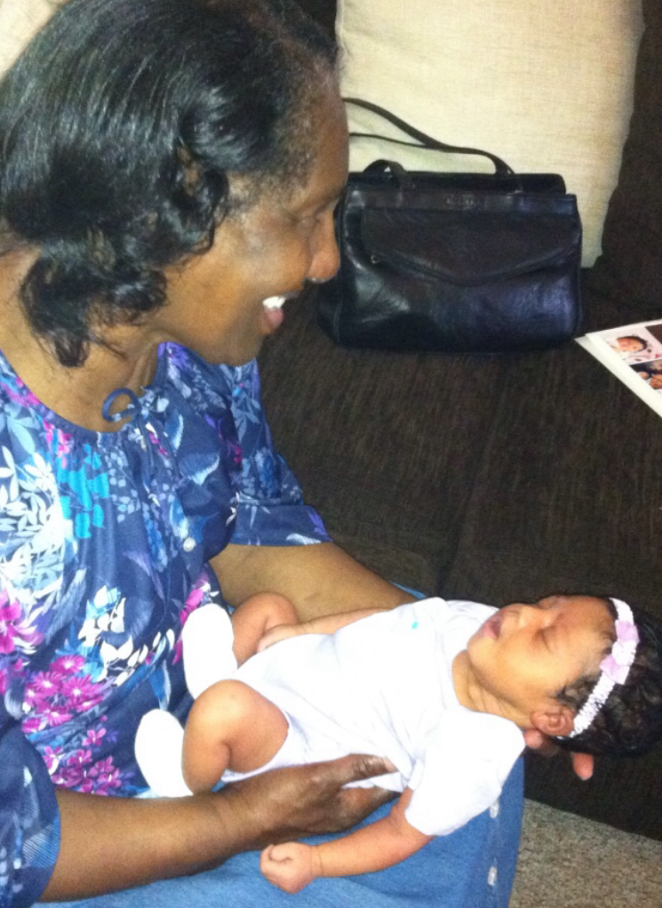 Woman seated on a sofa, smiling as she cradles a newborn baby dressed in white. A dark purse rests on the couch behind them.