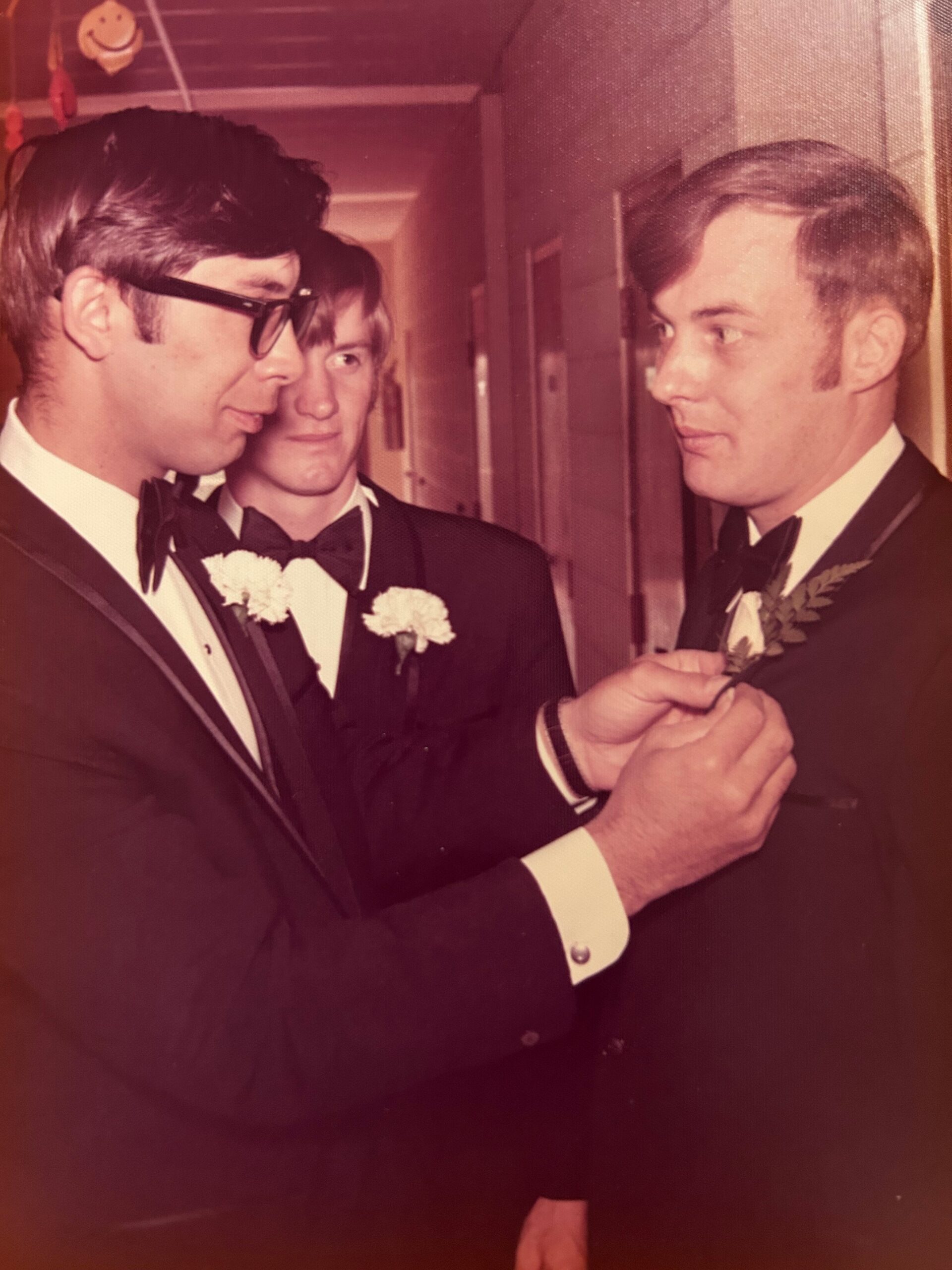 Three men in tuxedos at a formal event; one pins a boutonniere on another while two look on.