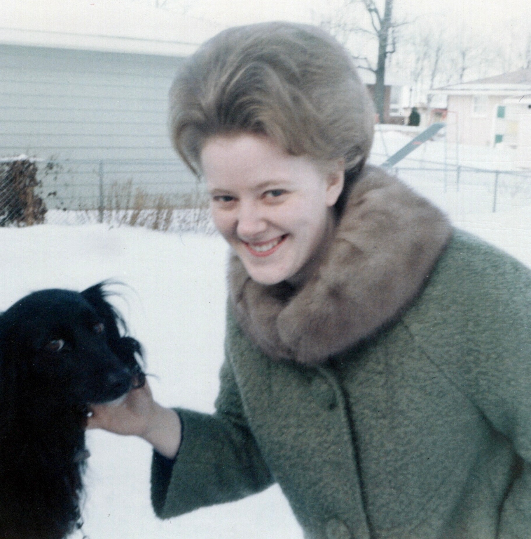 Smiling woman in a green fur-collared coat outdoors in snow, with a black dog partially visible in the foreground.