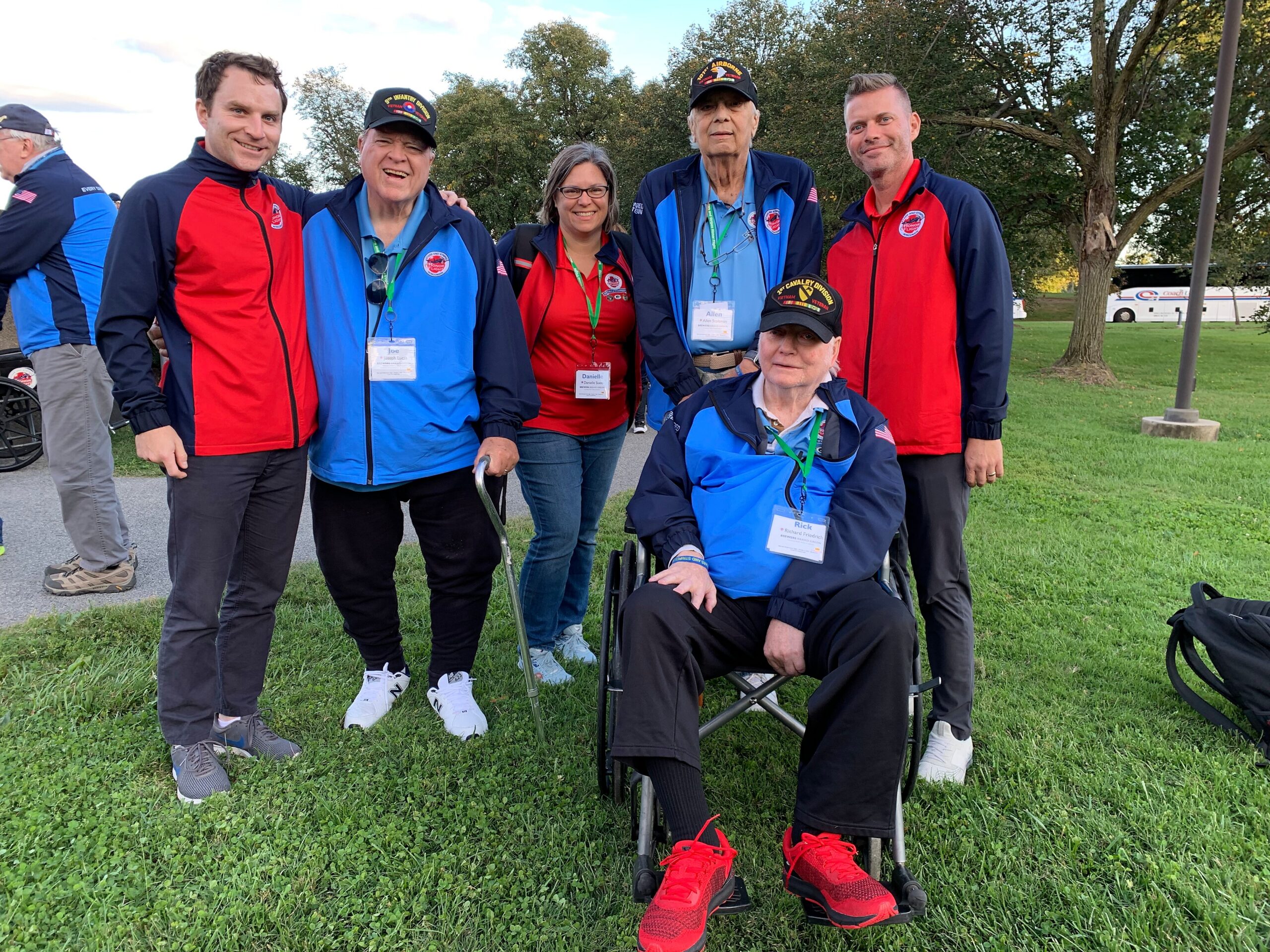 Group of veterans posing outdoors in red and blue jackets with badges, a person in a wheelchair at the front center, on a grassy area.
