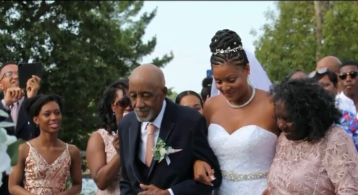 Bride in a white strapless gown walking arm-in-arm with an elderly man in a suit at an outdoor wedding, surrounded by guests.