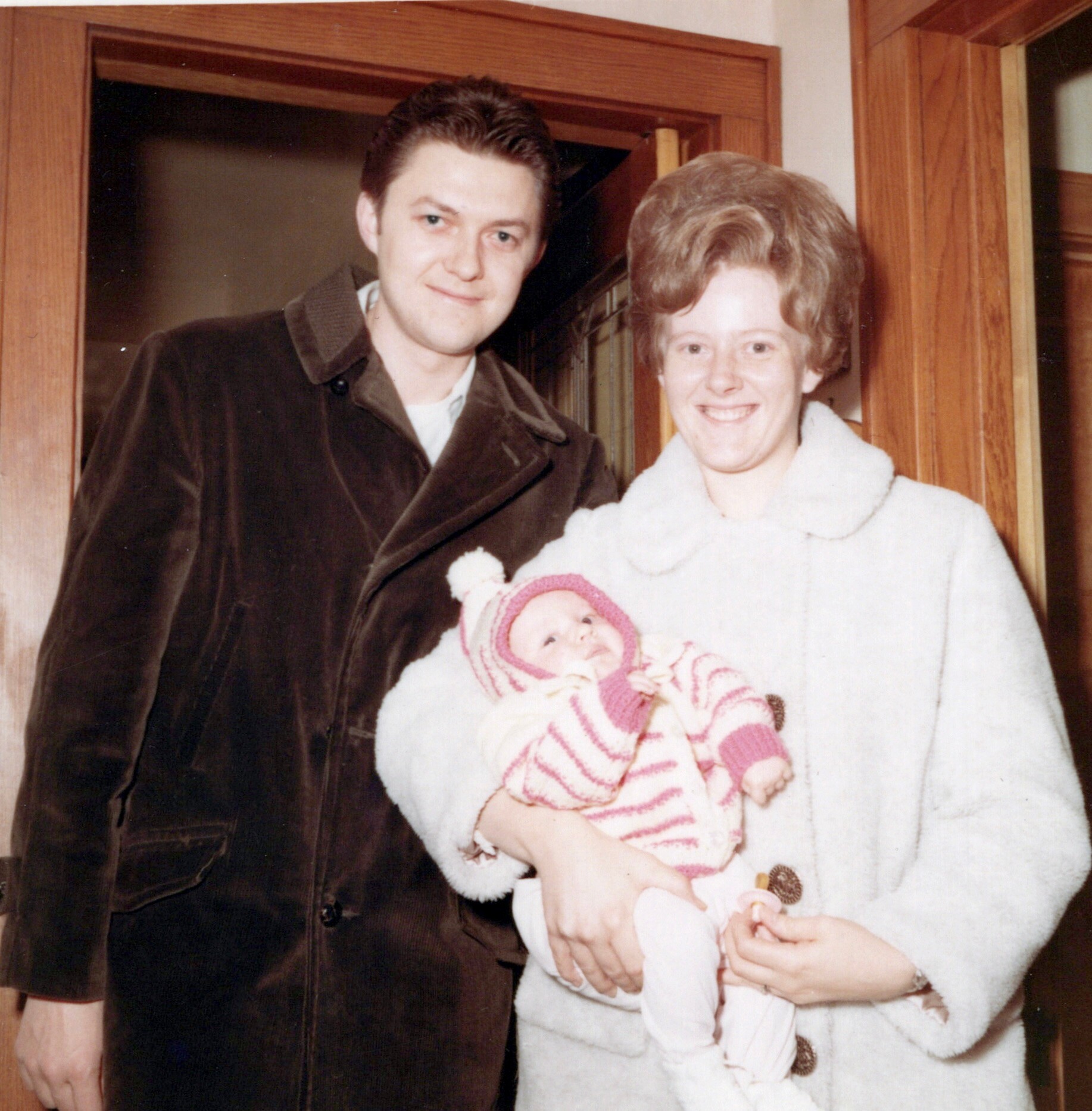 Man in brown jacket stands beside smiling woman holding a newborn baby wrapped in a pink-striped blanket inside a doorway.