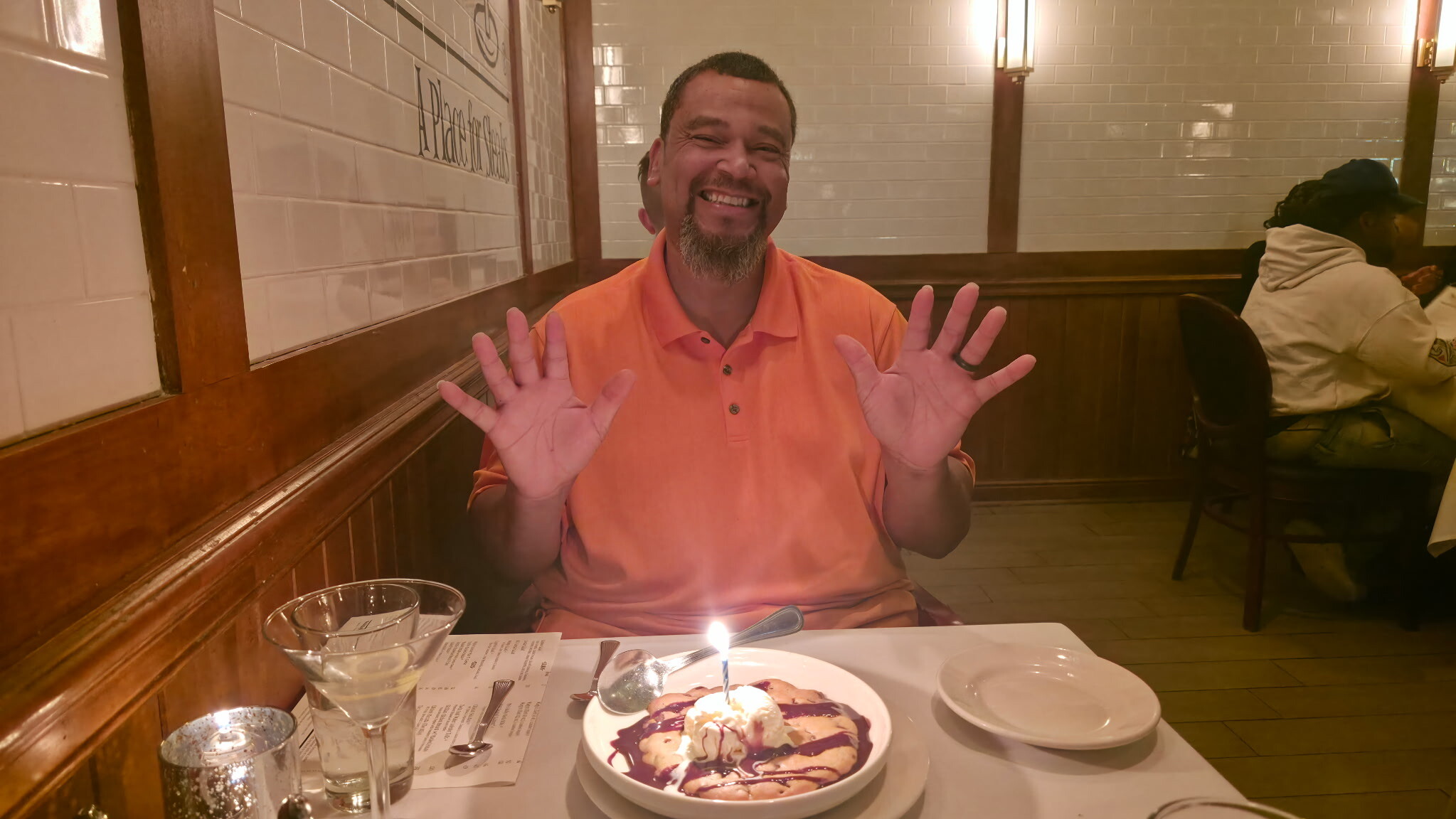 Smiling man in an orange polo raises both hands at a restaurant table with a small birthday cake and candle in front of him.