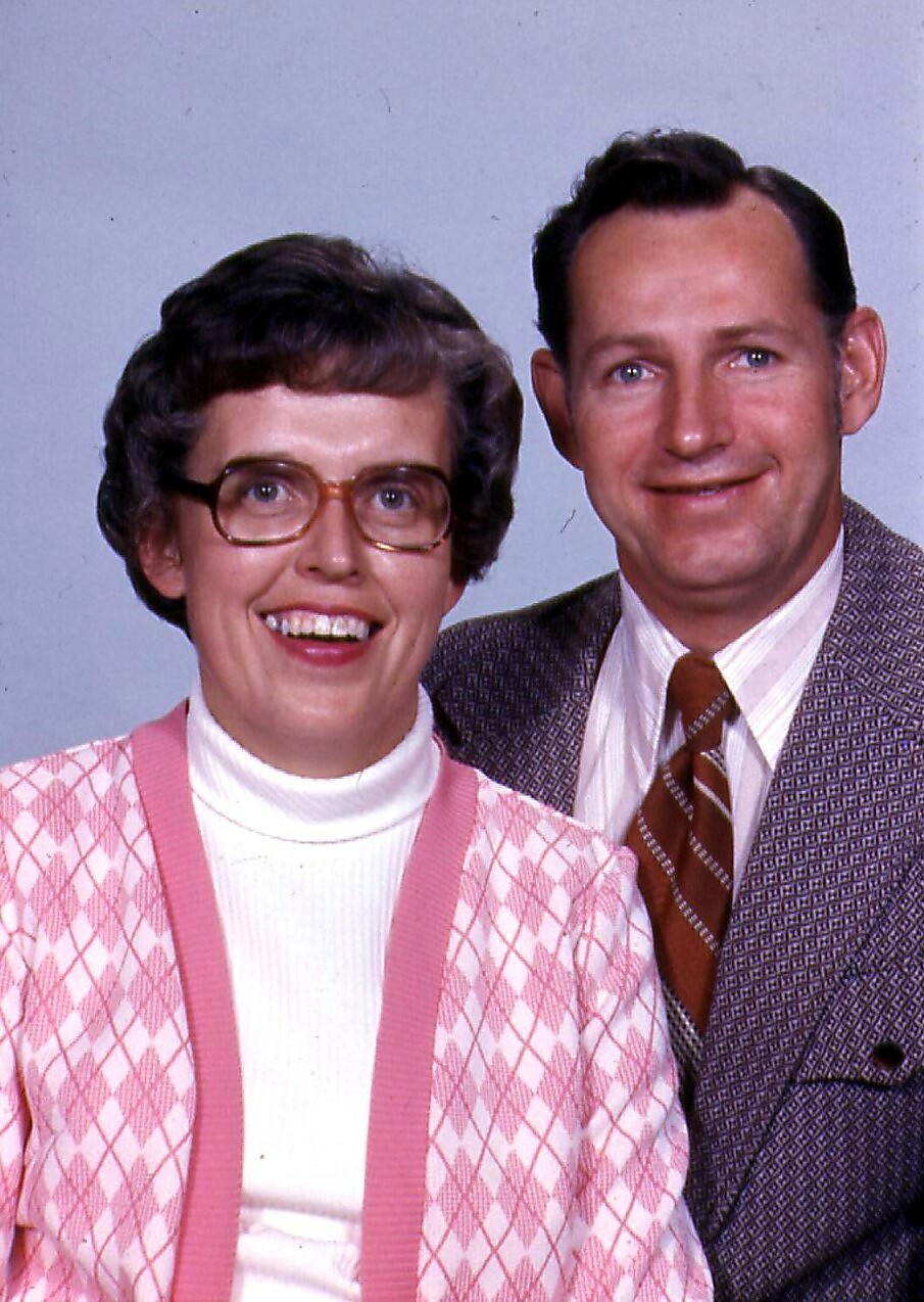 Smiling couple posing for a portrait: woman with short brown hair and glasses in a pink argyle cardigan, man in a brown tweed jacket and striped tie.