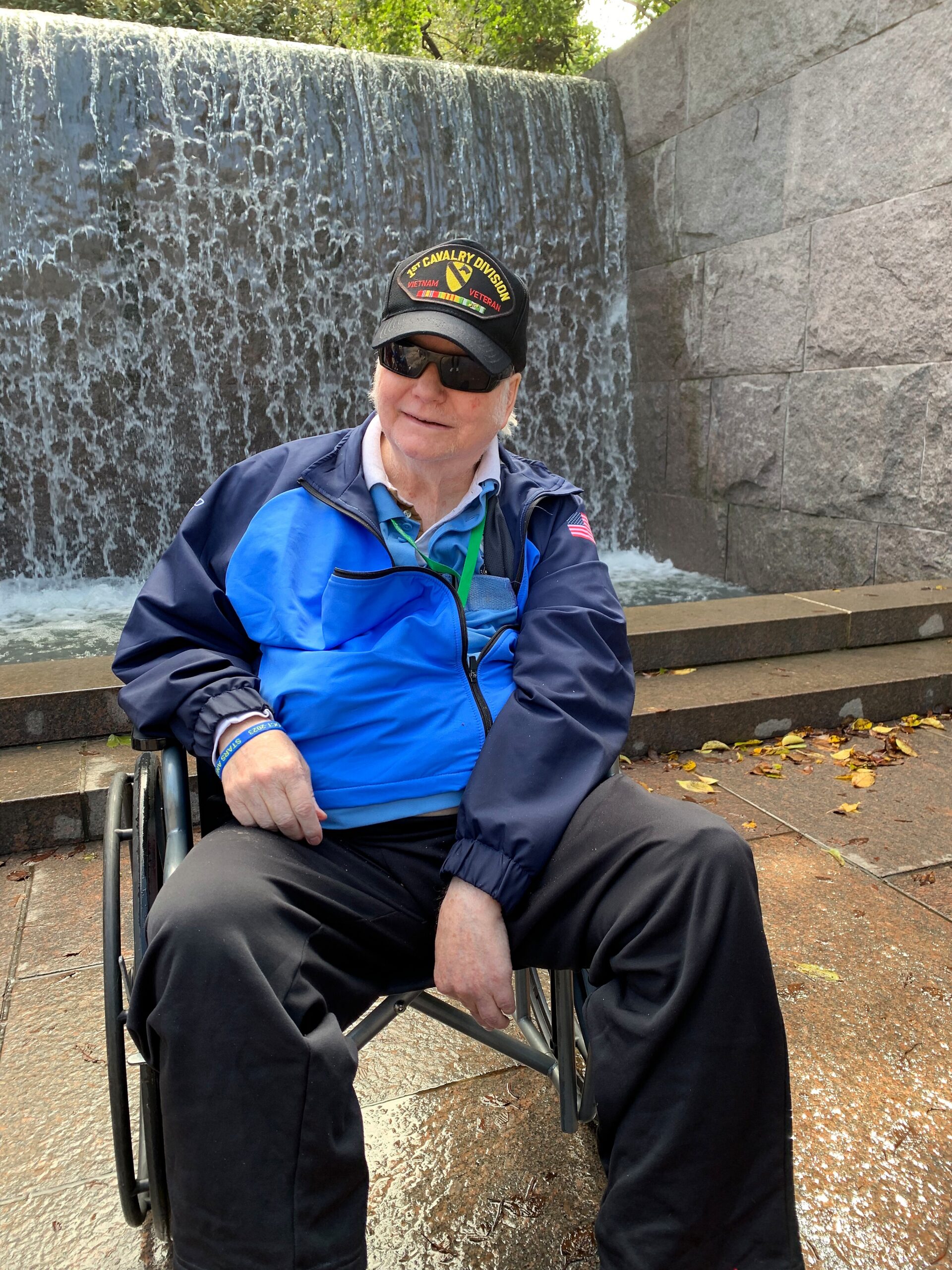 Man in a wheelchair wearing a blue and navy jacket and a black cap with a Vietnam veteran patch, outdoors near a cascading water feature.