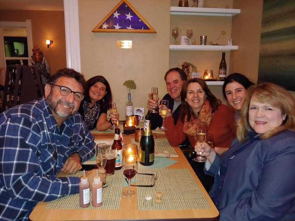 Seven adults around a dining table toasting with glasses of wine/champagne in a cozy restaurant, candles lit and bottles on the table.