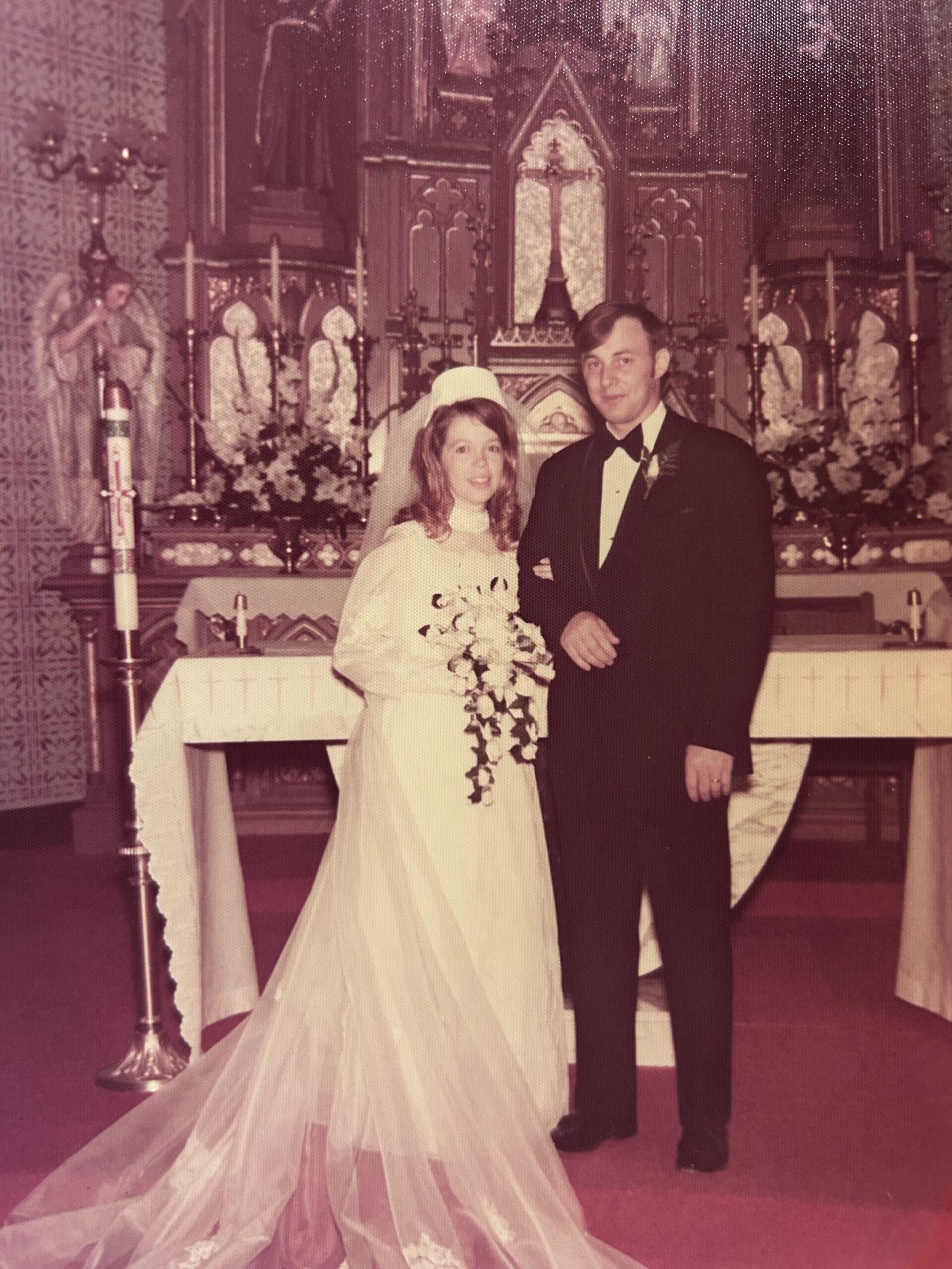 Bride in a wedding dress with bouquet and veil standing beside a groom in a tuxedo at the church altar.