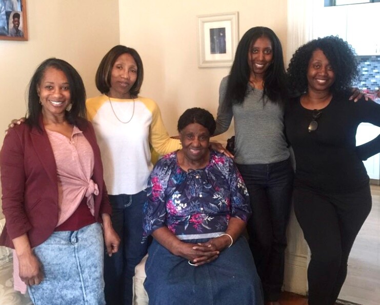 Older woman seated in a blue floral top among four standing women in a cozy indoor setting, all smiling at the camera.