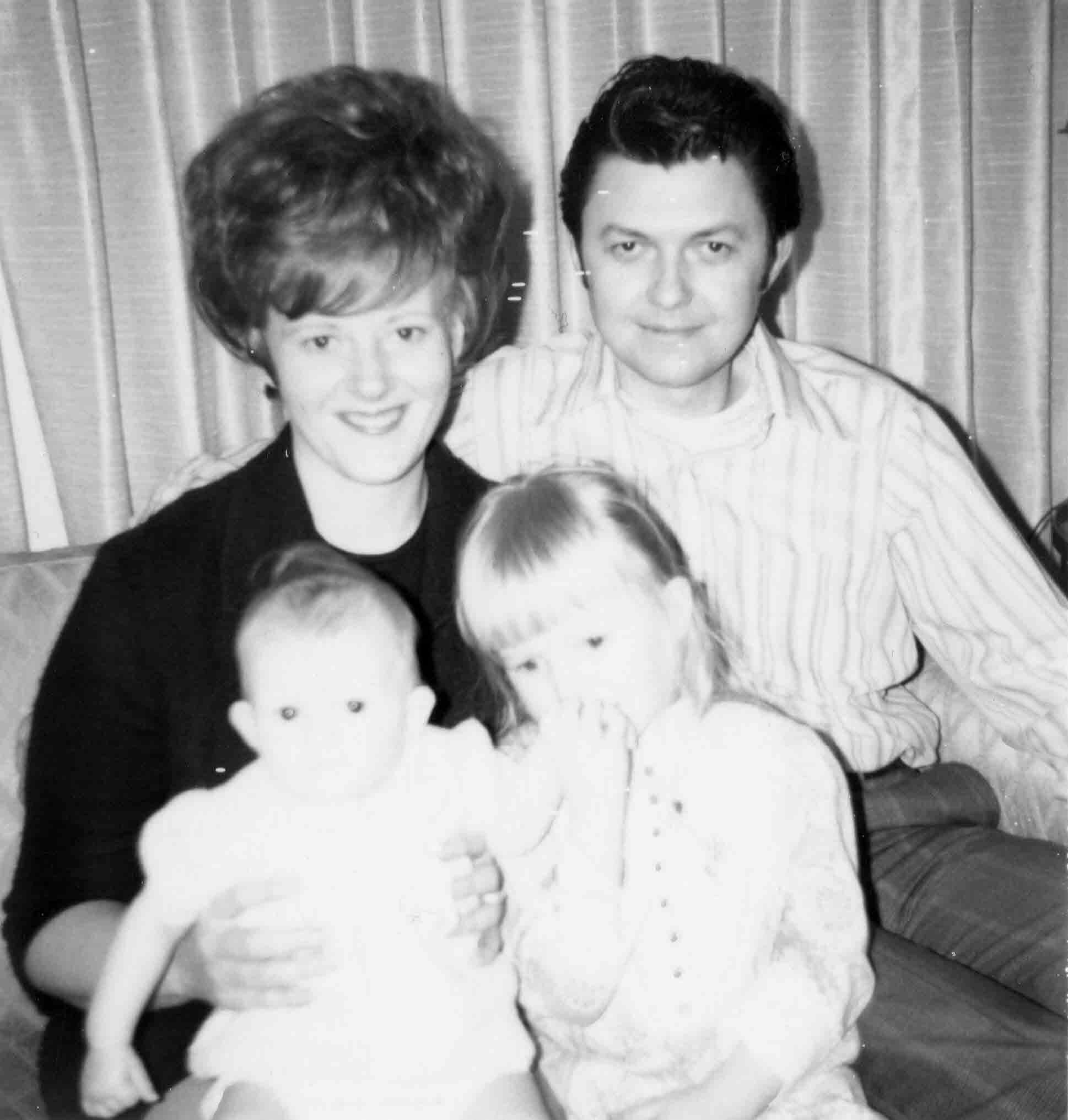 Black-and-white family photo: a smiling woman and man with two babies in front, seated on a couch with curtains in the background.